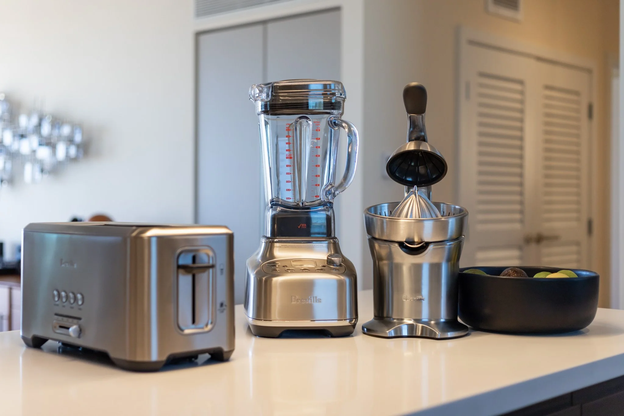 A kitchen countertop with a stainless steel toaster, a blender, a manual citrus juicer, and a black bowl filled with assorted fruit.