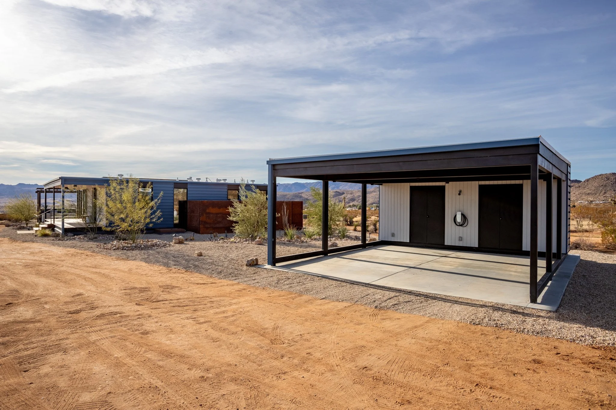 A modern, single-story house with a detached covered carport in a desert landscape, with mountains in the background and a partly cloudy sky.