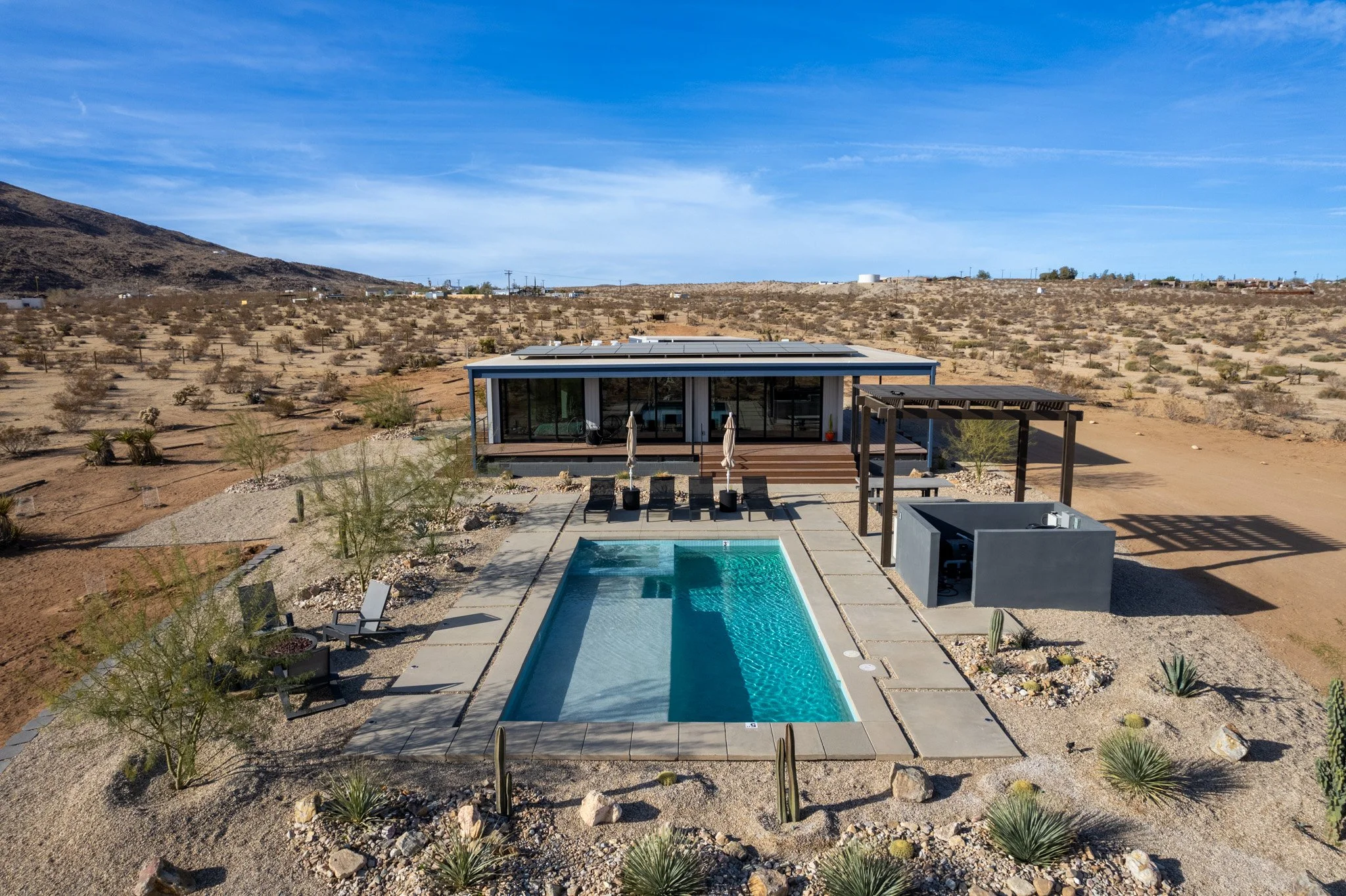 Modern house with a backyard pool in a desert landscape, featuring lounge chairs and a shaded patio area under a pergola.