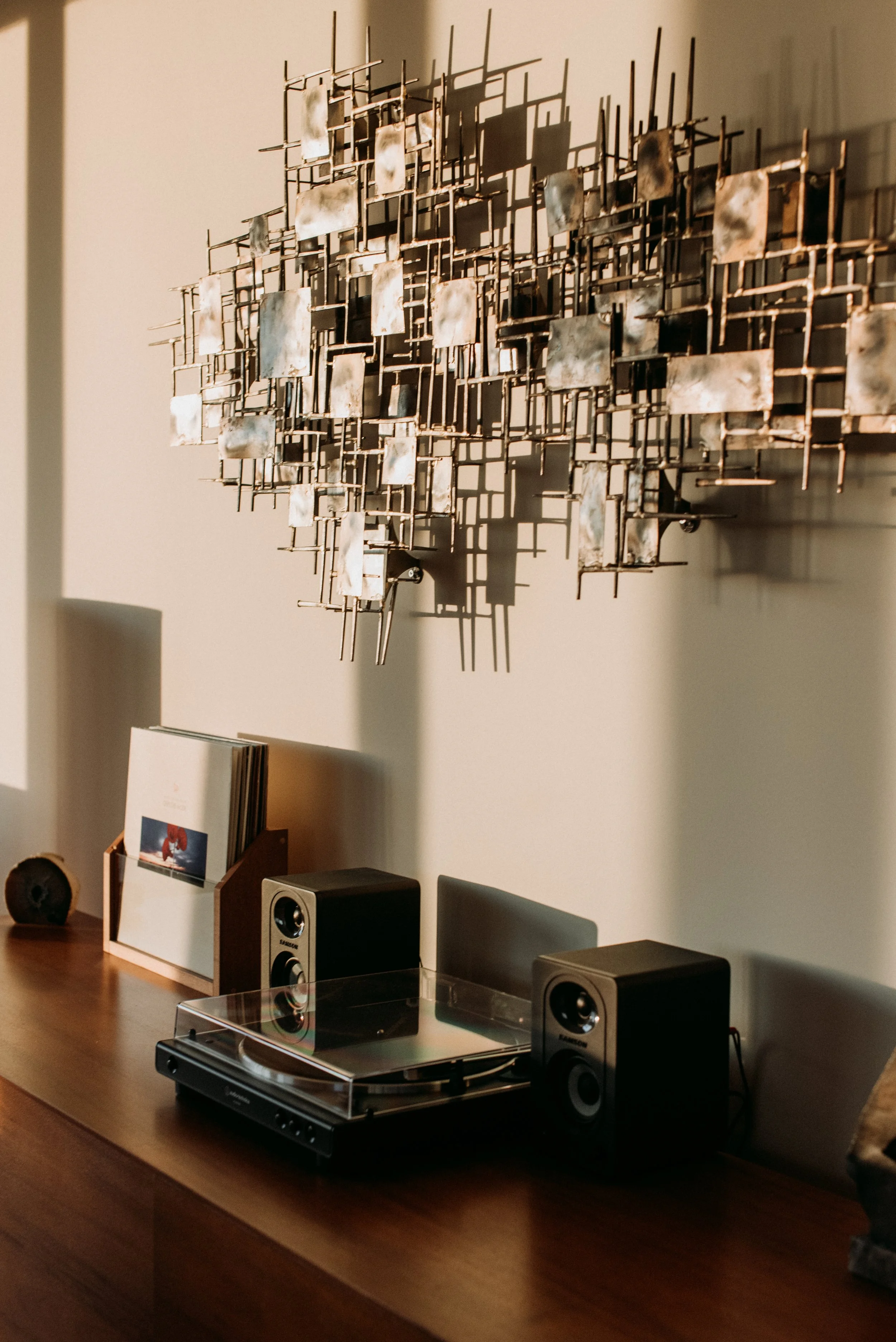 A wooden sideboard with a Sony turntable, two speakers, and a small wooden rack with vinyl records. There is an abstract metal wall sculpture hanging above the sideboard, and sunlight casting shadows on the wall.