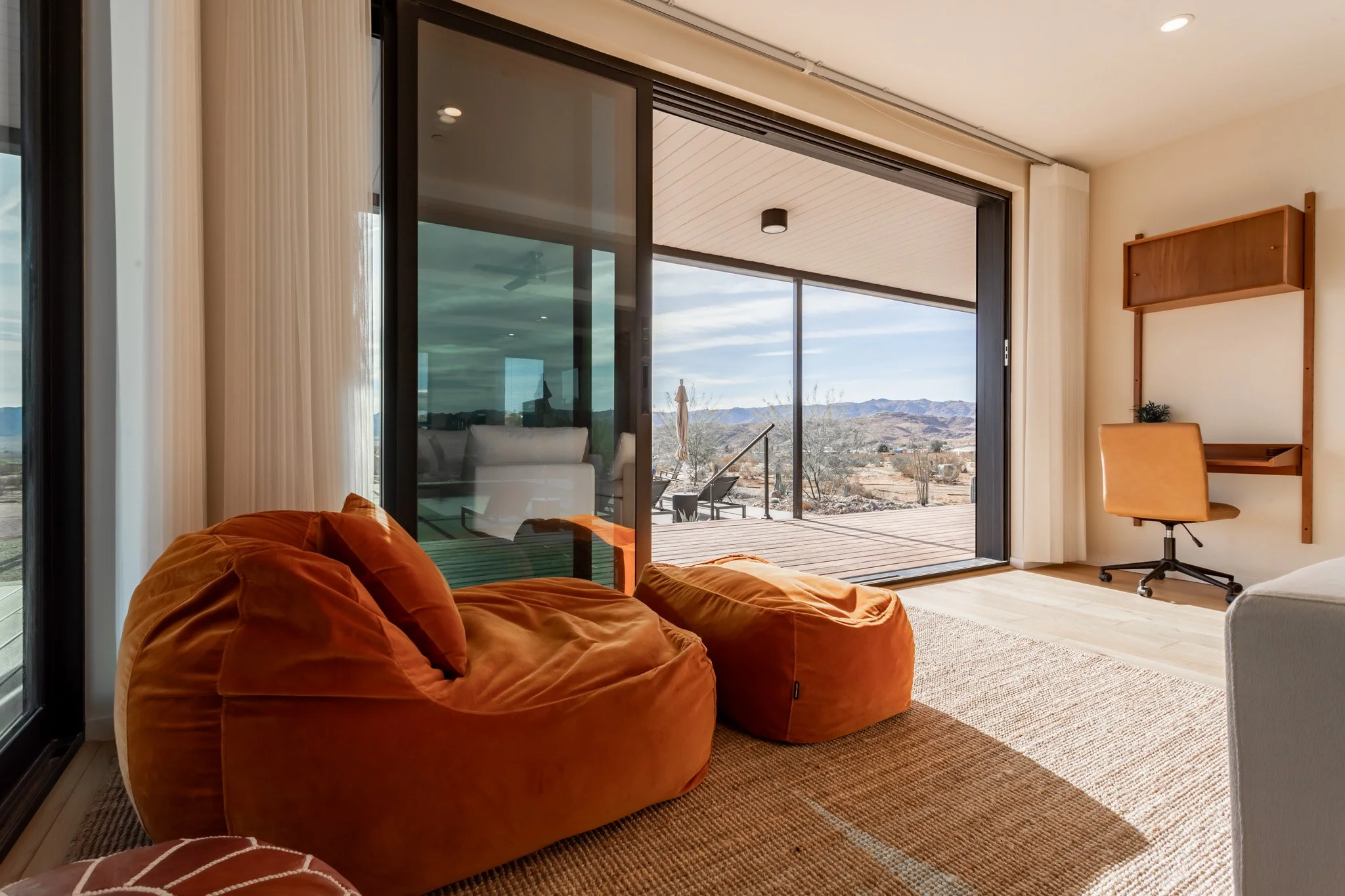 Living room with large glass sliding doors leading to a desert landscape outdoor patio, featuring orange bean bag chairs, a small desk with a chair, and mountains in the distance.