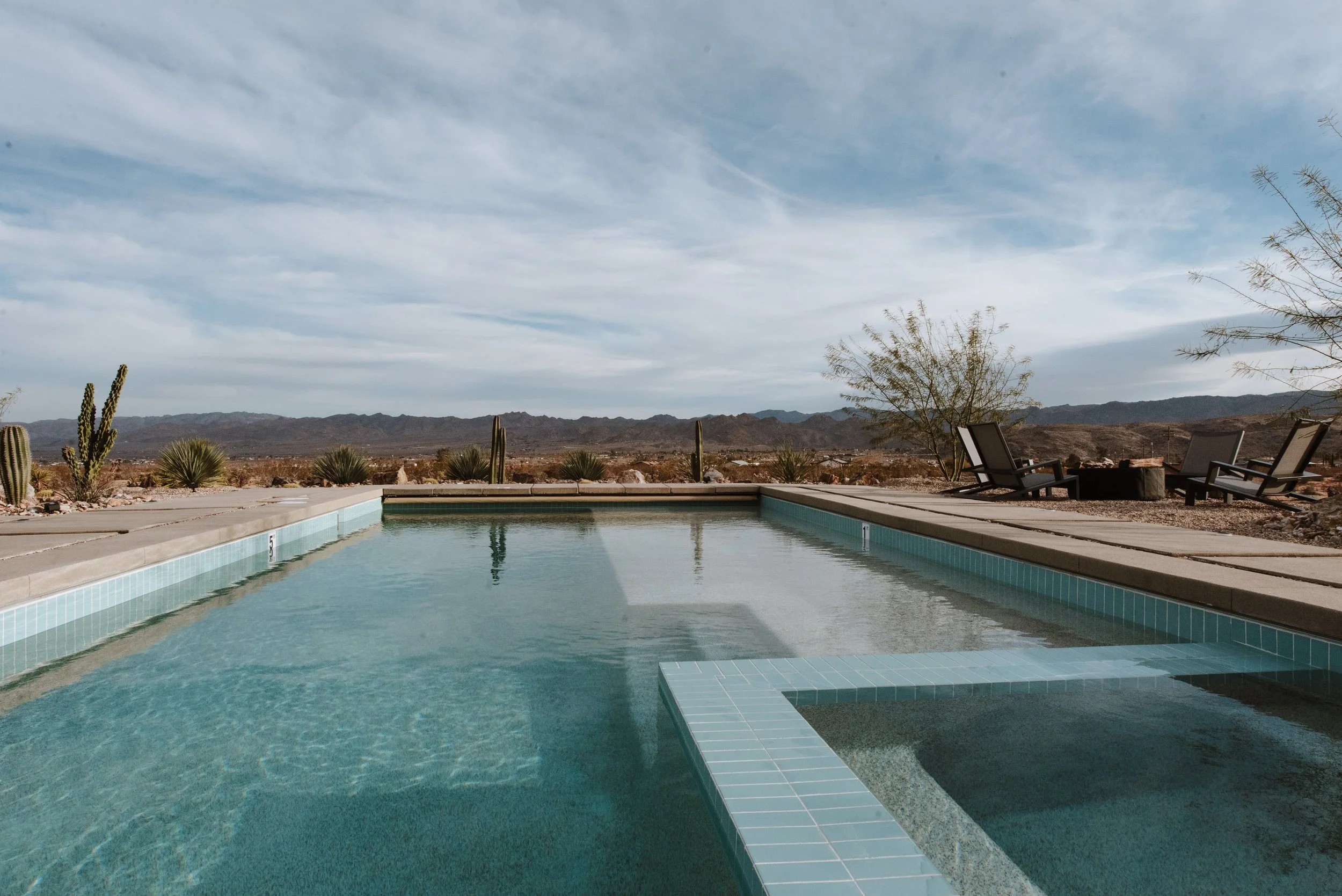 An outdoor swimming pool with desert landscape and mountains in the background, plus lounge chairs and a fire pit on the patio.