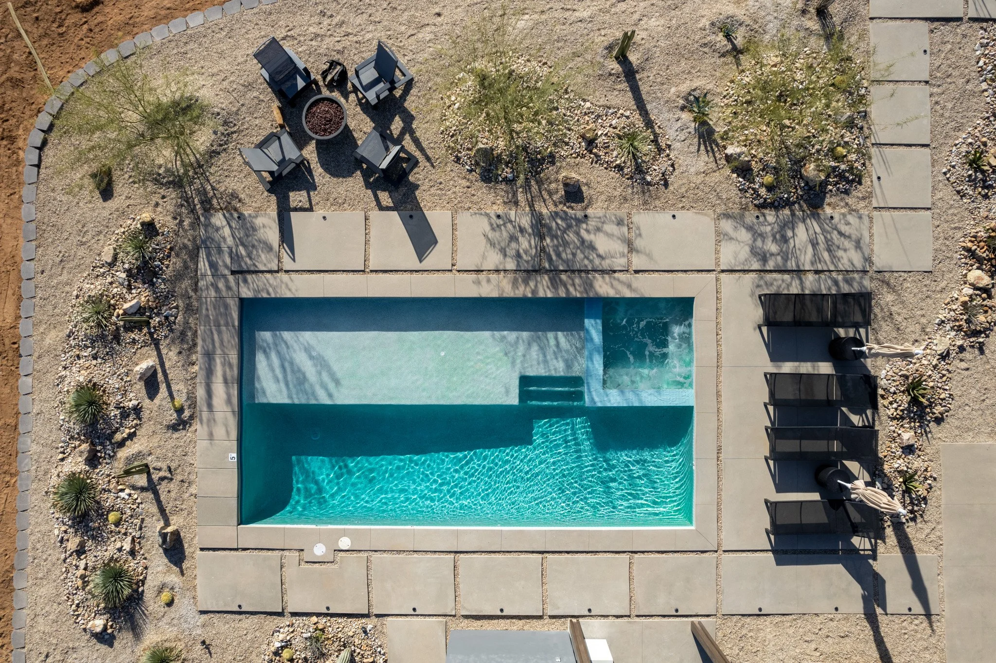 Aerial view of a rectangular swimming pool with built-in steps, surrounded by concrete pavers, with outdoor seating, trees, and desert landscaping.