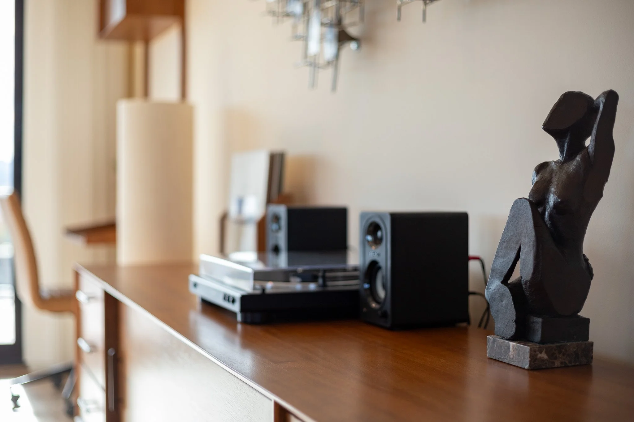 A wooden sideboard with a decorative sculpture, a vintage record player, and speakers, in a room with a beige wall and framed pictures in the background.
