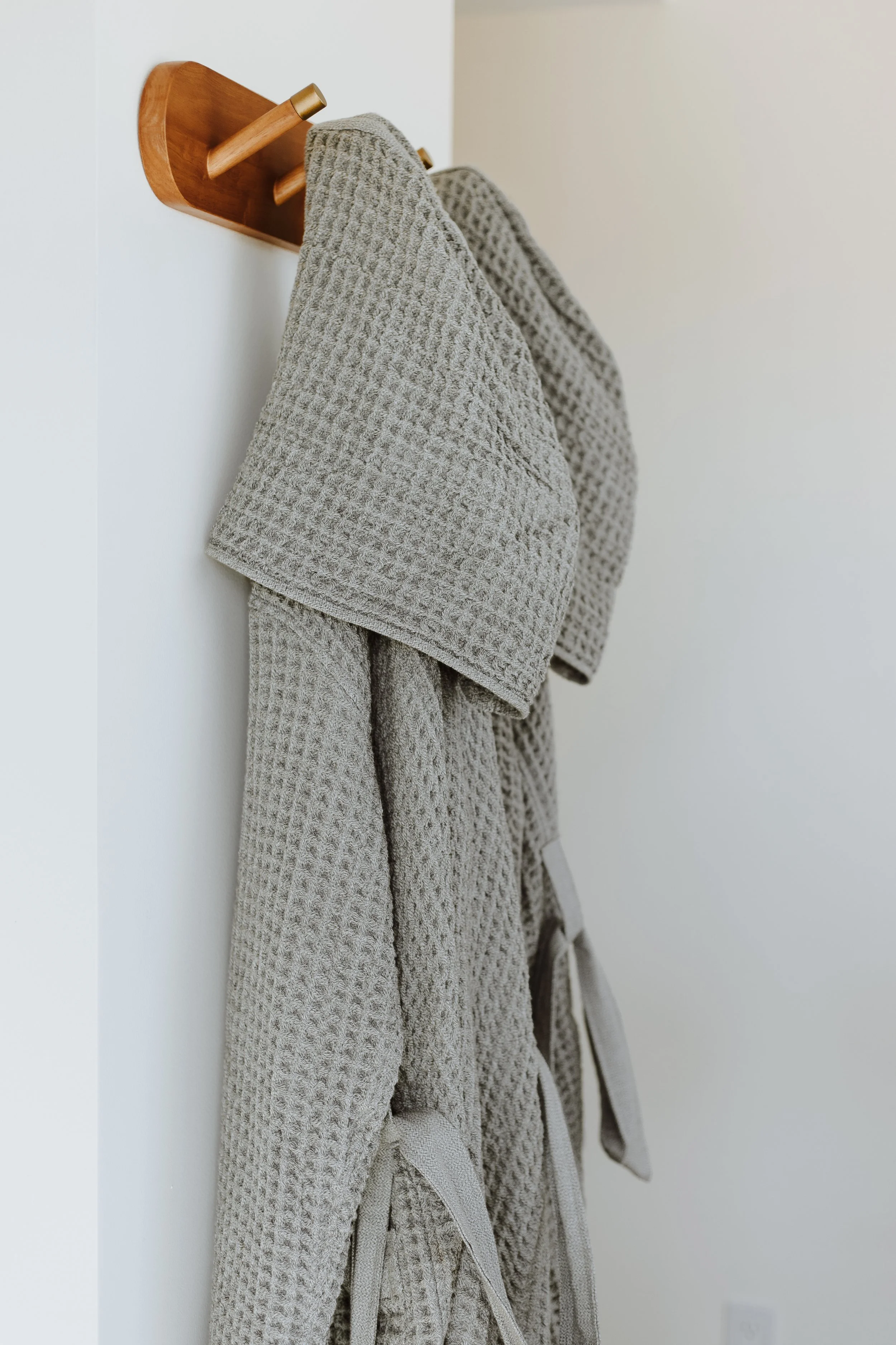 Gray waffle-knit bathrobe hanging on a wooden wall hook in a white room.