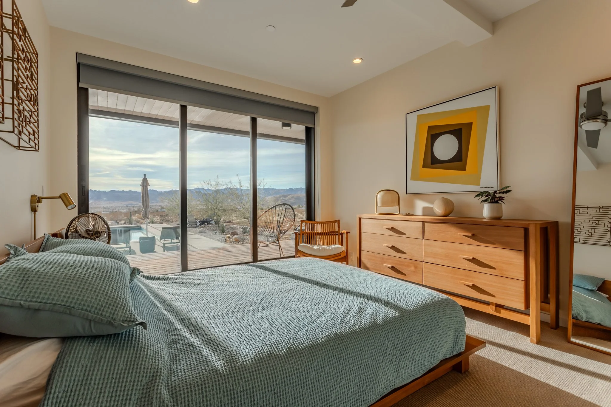 Bedroom with a large sliding glass door opening to a desert landscape and mountain view, a made bed with green bedding, a wooden dresser with abstract art, decorative objects, and a chair, with sunlight casting shadows inside.