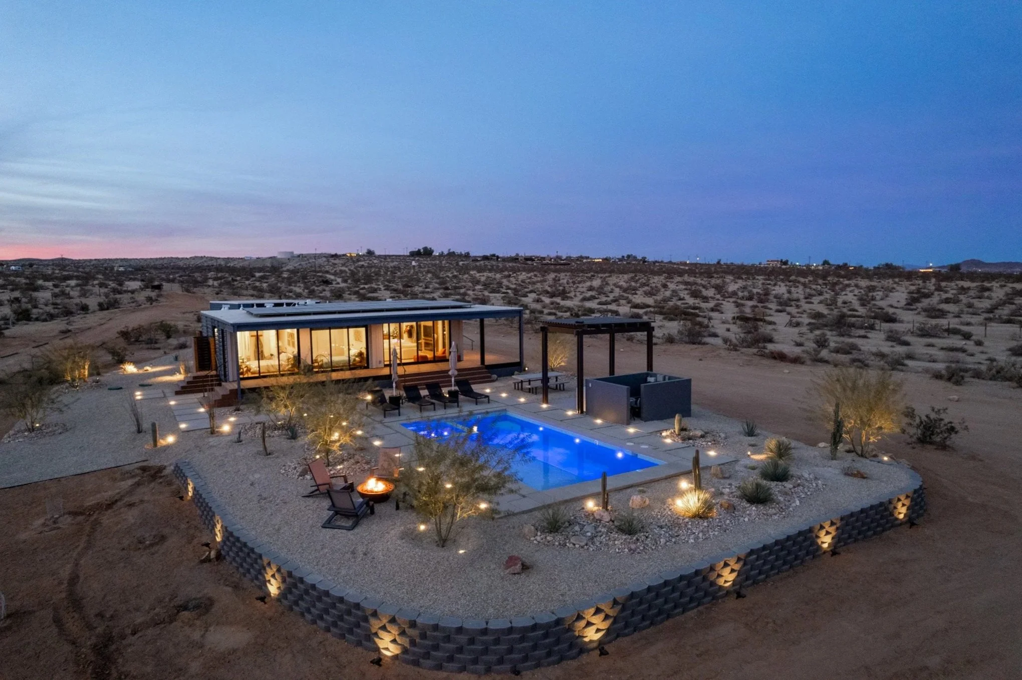 A modern single-story house in a desert landscape at dusk with a lit swimming pool, outdoor seating, and ambient lighting.