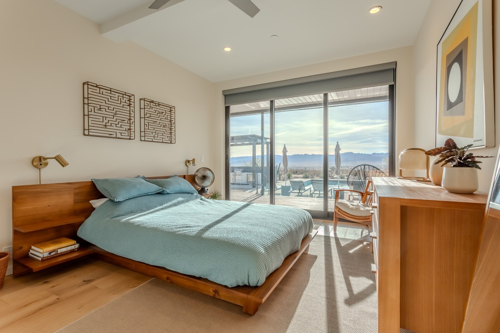Bedroom with wooden bed, light blue bedding, and a view of an outdoor patio through sliding glass doors, with mountains in the background.