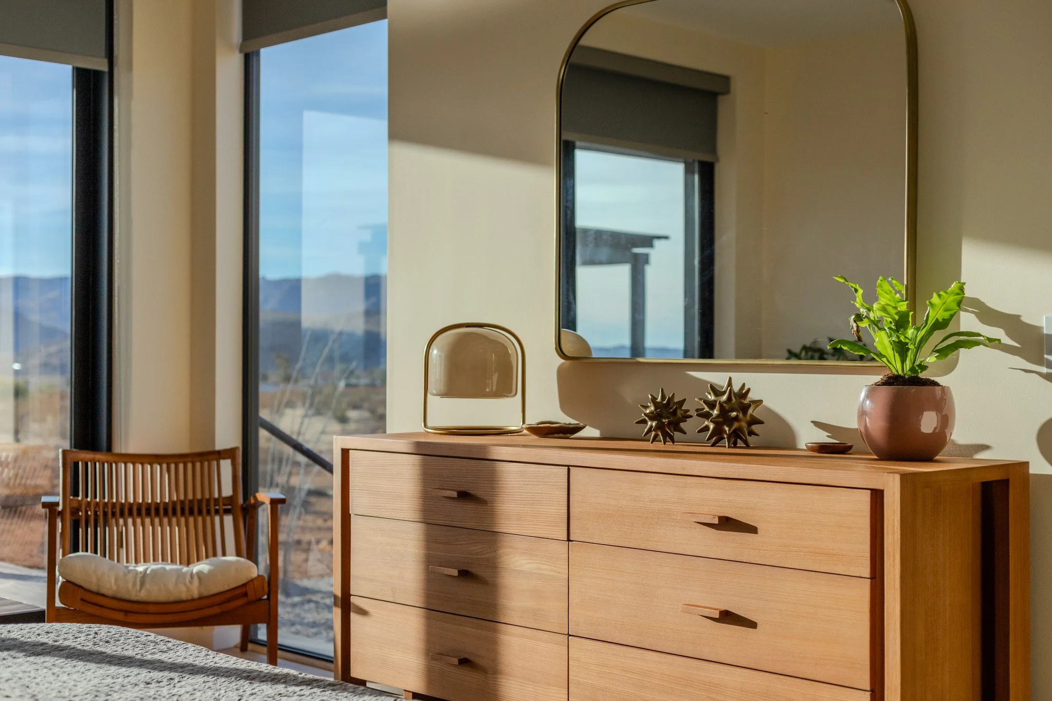 Interior of a bedroom with a wooden dresser, a large mirror, a potted plant, two decorative objects, a table lamp, a wooden chair with a cushion, and large windows showing an outdoor landscape with mountains.
