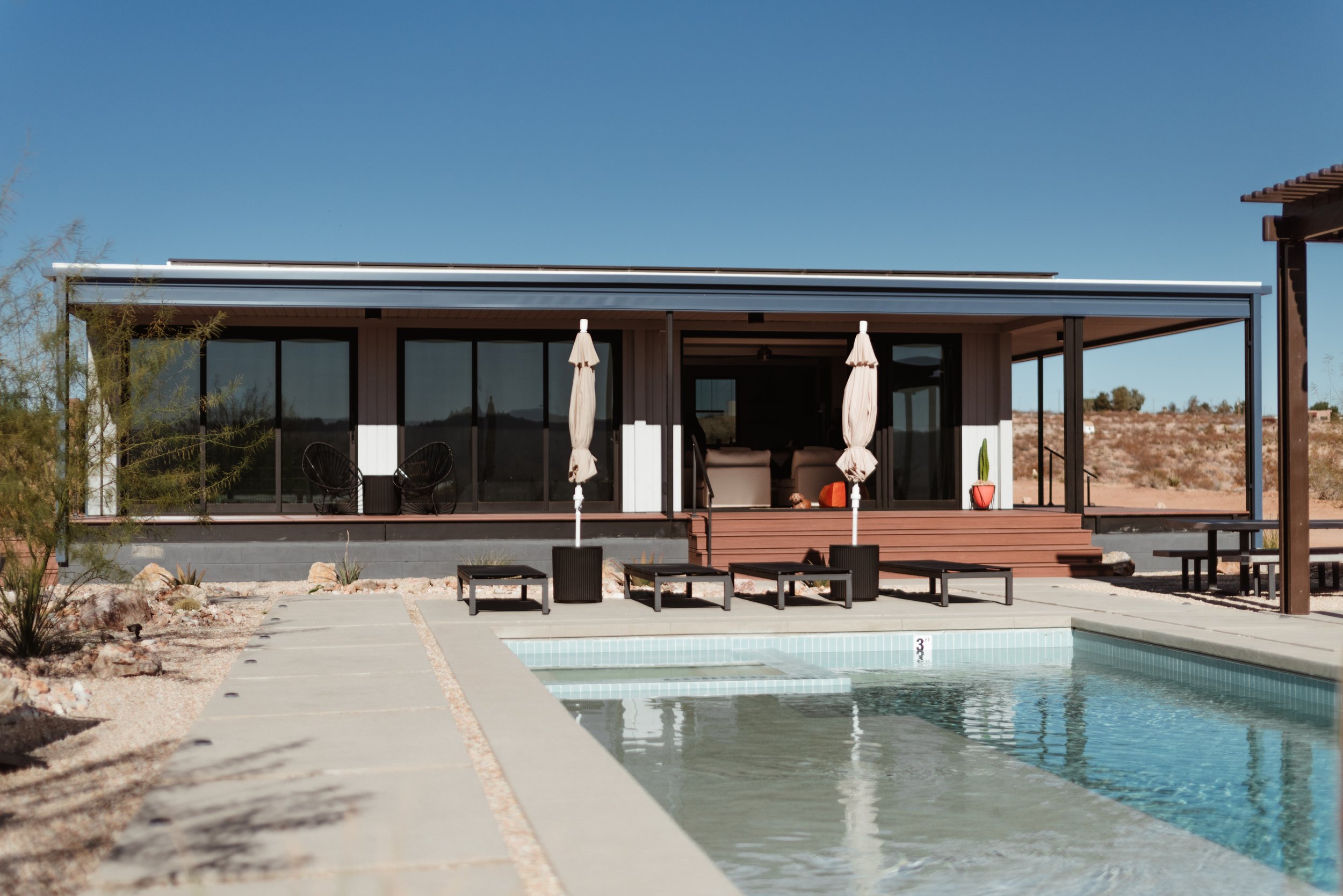 Modern house with large glass sliding doors, outdoor seating, umbrellas, potted plants, and a backyard swimming pool under a clear blue sky.