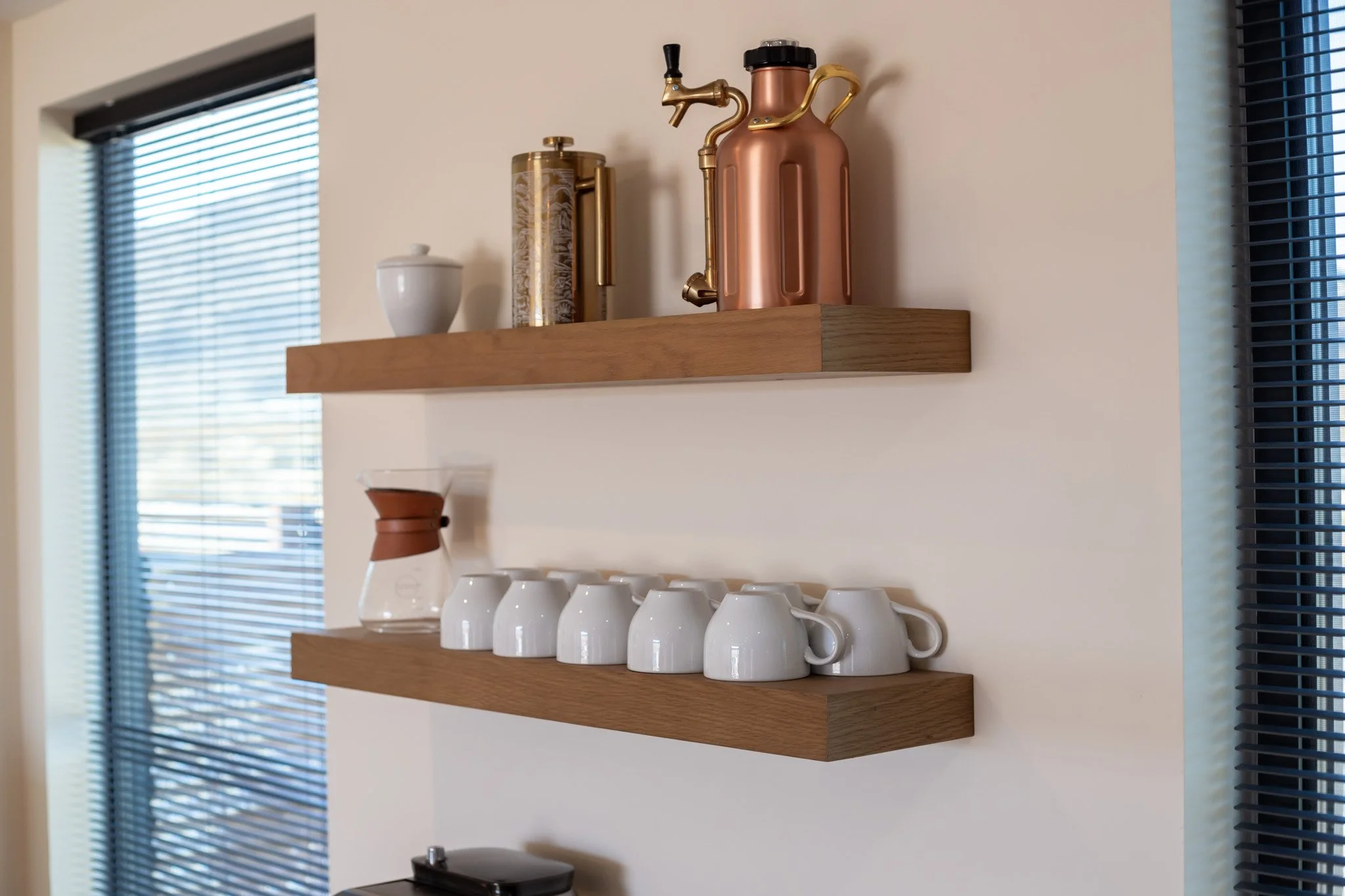 Two wooden shelves on a wall, top shelf has a white container, a gold and silver thermos, and a copper-colored siphon coffee maker. The bottom shelf has a glass Chemex coffee maker and six white coffee cups with handles.