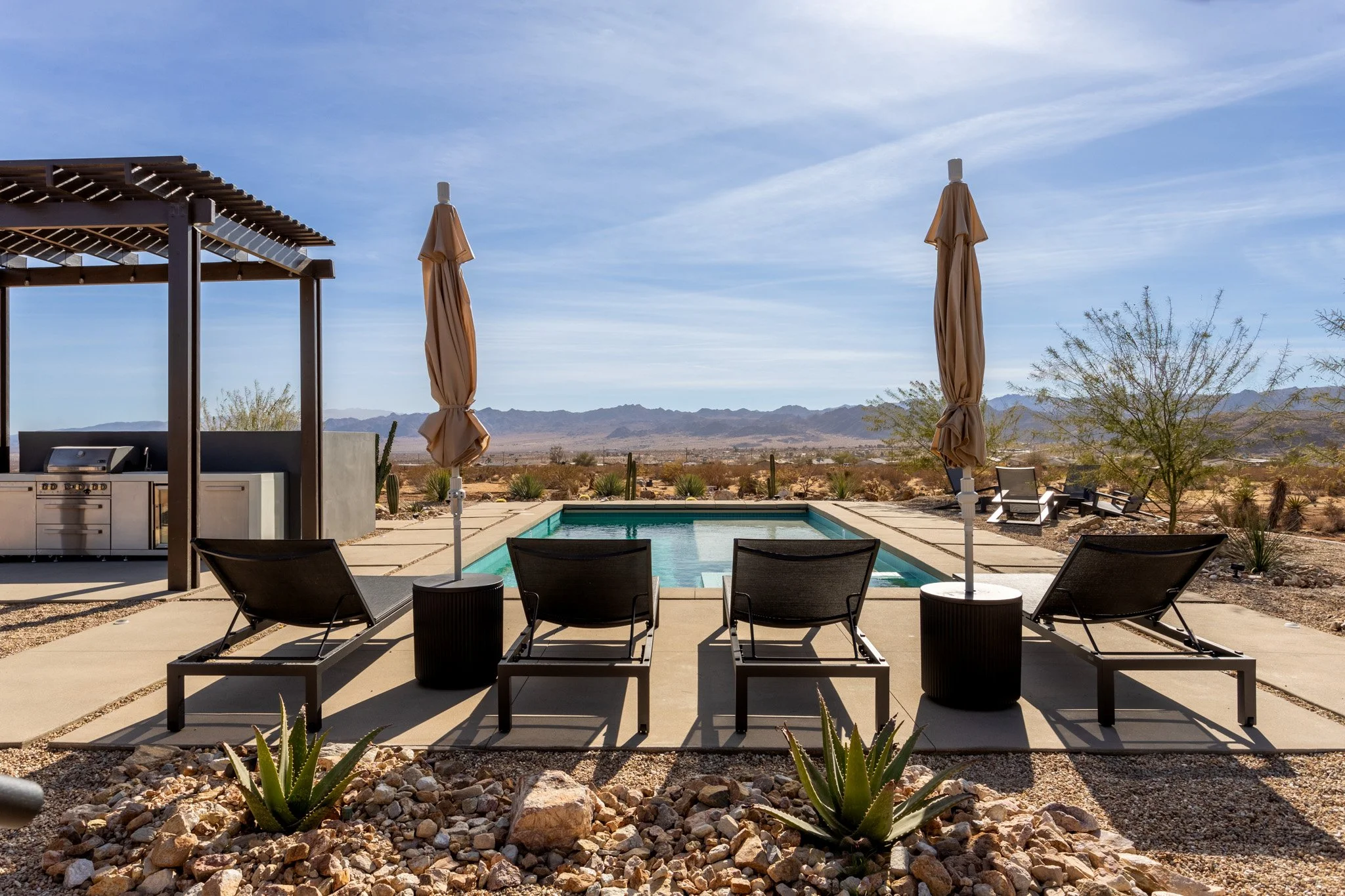 A backyard with a swimming pool, lounge chairs, umbrellas, and desert plants with mountains in the background under a blue sky.