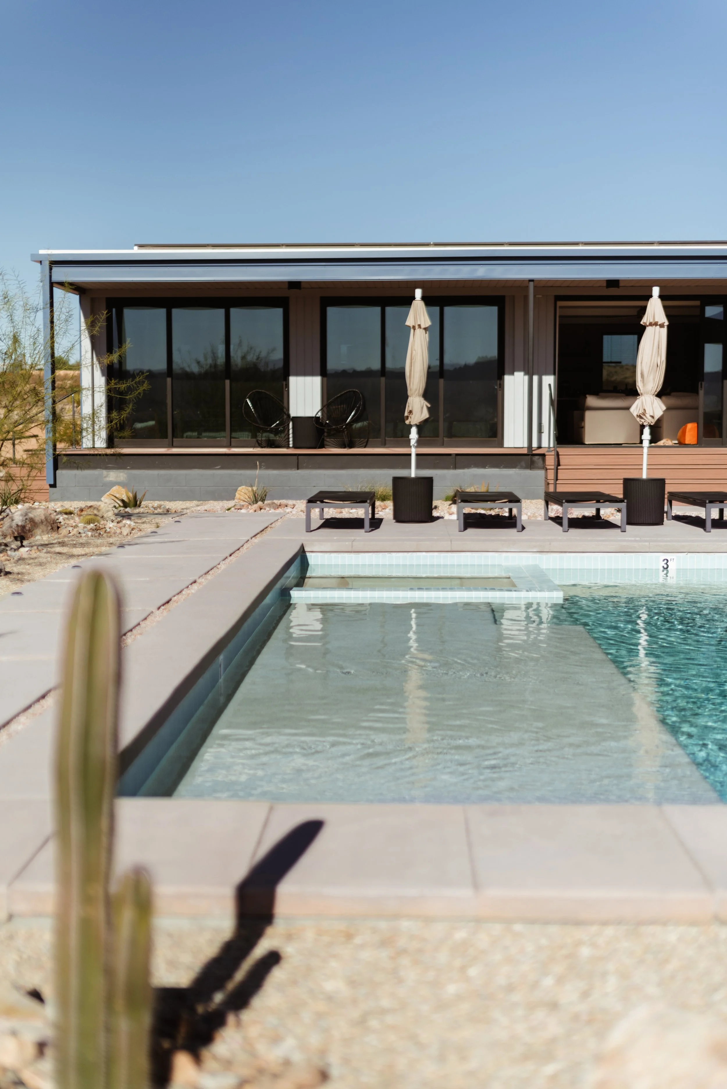Modern house with large glass doors, outdoor swimming pool with steps, lounge chairs, and umbrellas, against clear blue sky.