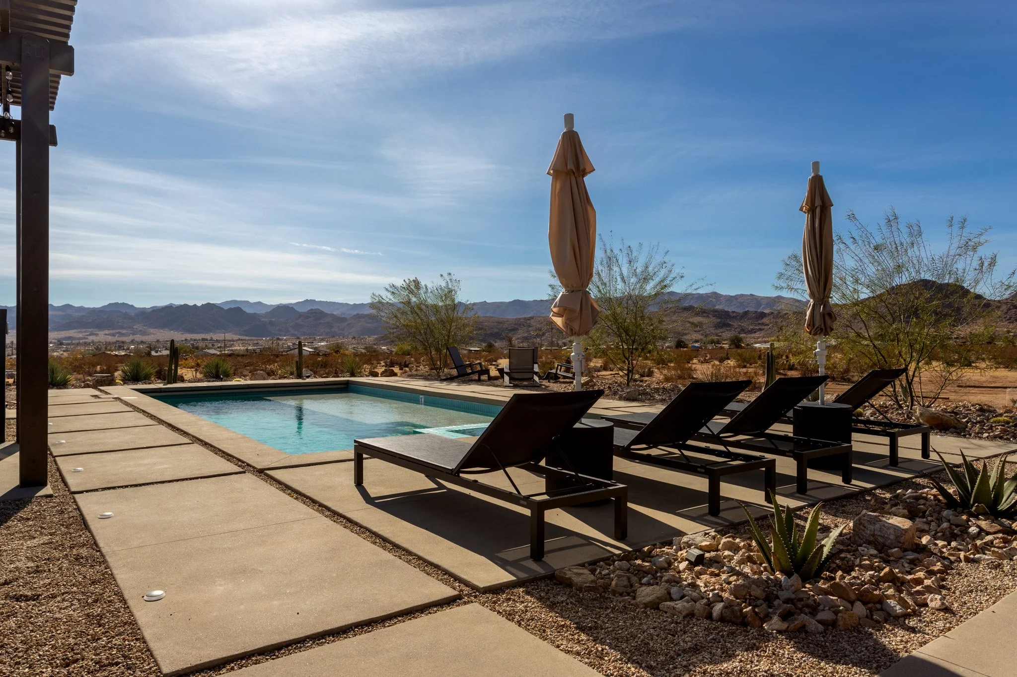 Desert landscape with swimming pool, lounge chairs, and umbrellas, with mountains in the distance under a clear blue sky.