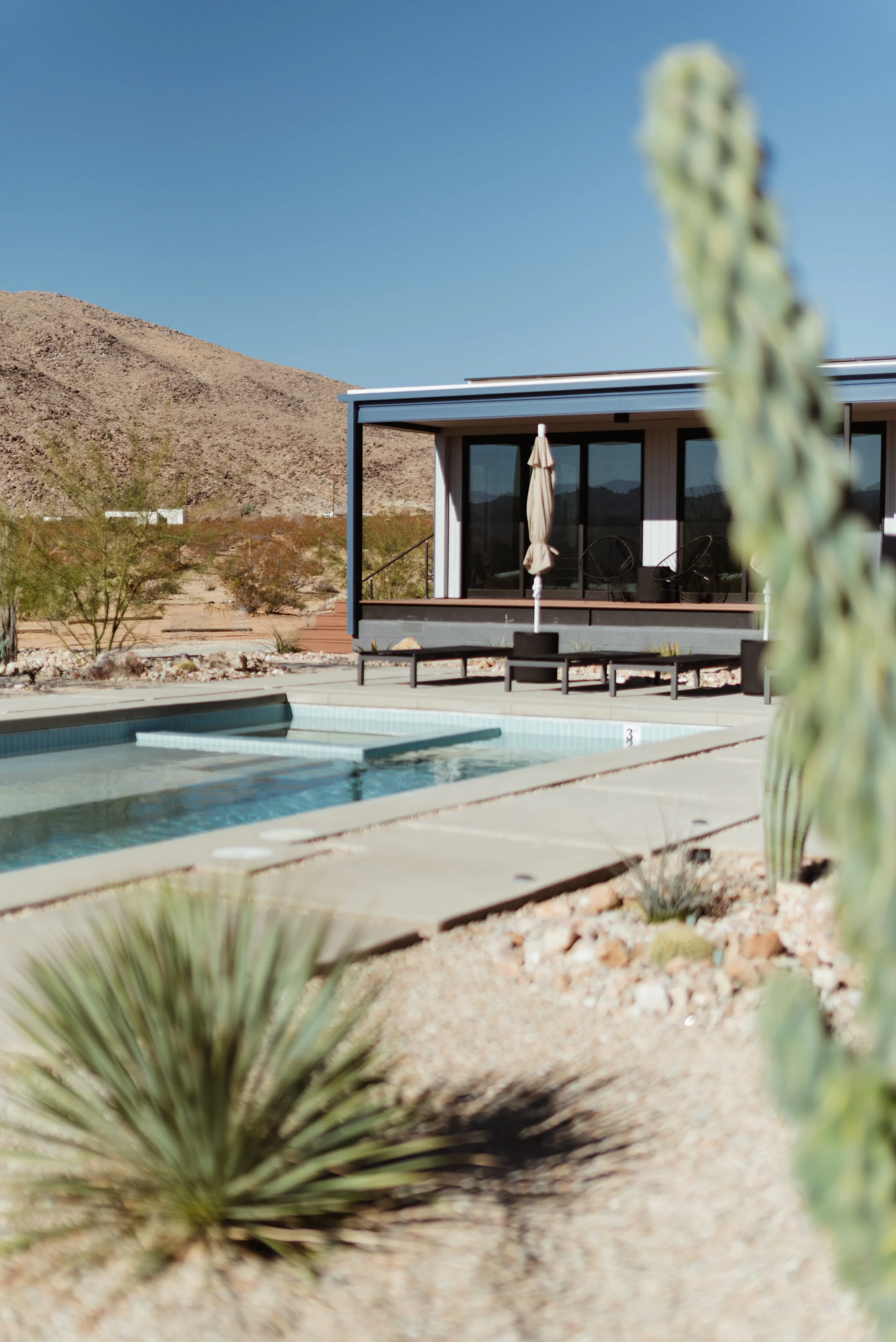 A desert landscape with a modern house, a swimming pool, desert plants, and mountains in the background under a clear blue sky.