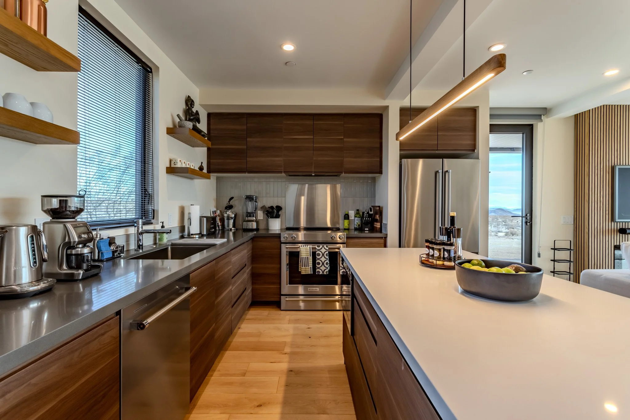 Modern kitchen with wooden cabinets, stainless steel appliances, a large island with a bowl of fruit, and a window showing an outdoor landscape.