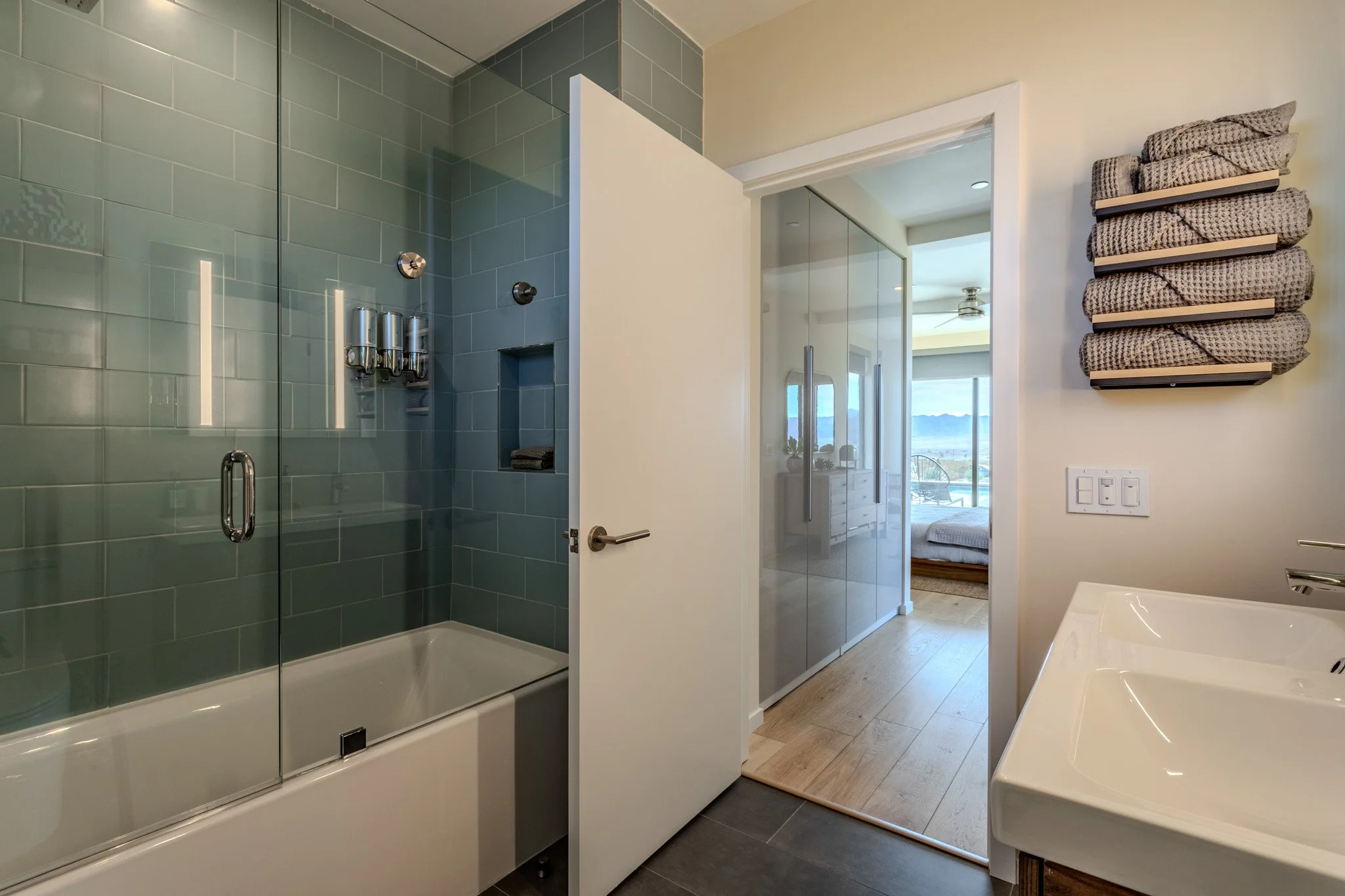 Modern bathroom with a glass-enclosed shower, white double sink vanity, countertop towels on a wall shelf, and a view of a bedroom with large windows and a mountain landscape.