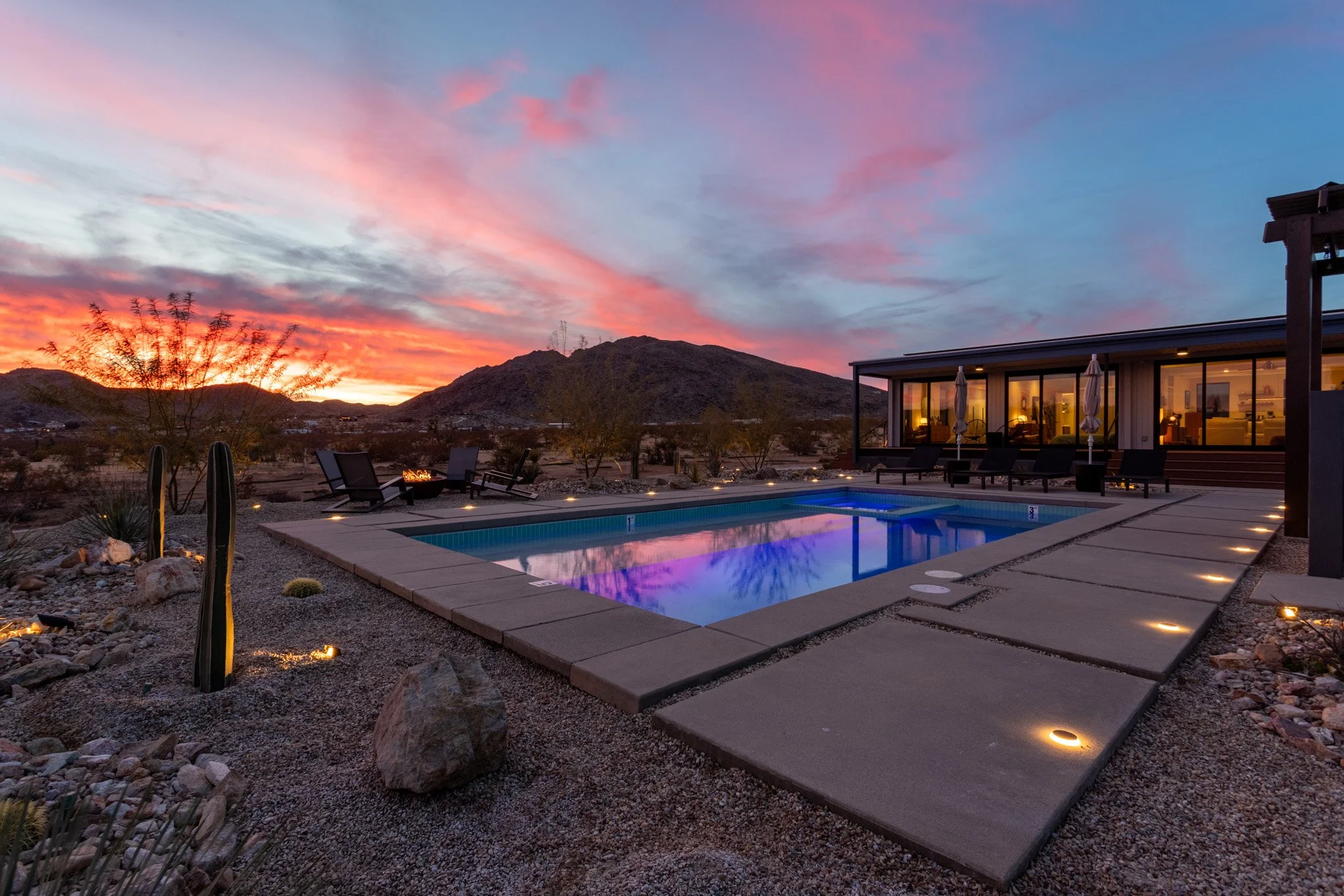 A nighttime view of a backyard with a lit swimming pool, surrounded by outdoor lounge chairs, desert plants, and a modern house with large glass windows, with a colorful sunset sky in the background and mountains in the distance.