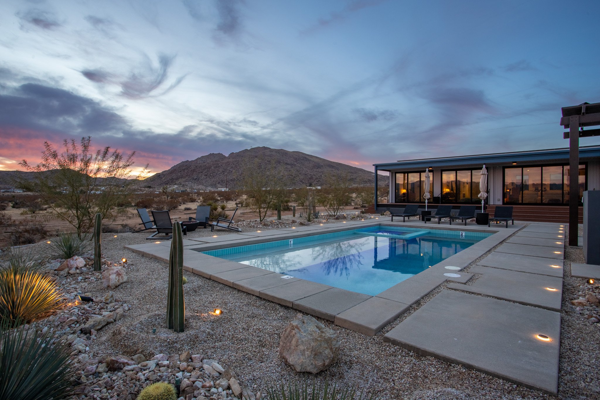 Desert landscape with a modern outdoor pool area during sunset, featuring lounge chairs, umbrellas, and a building with large glass windows in the background.