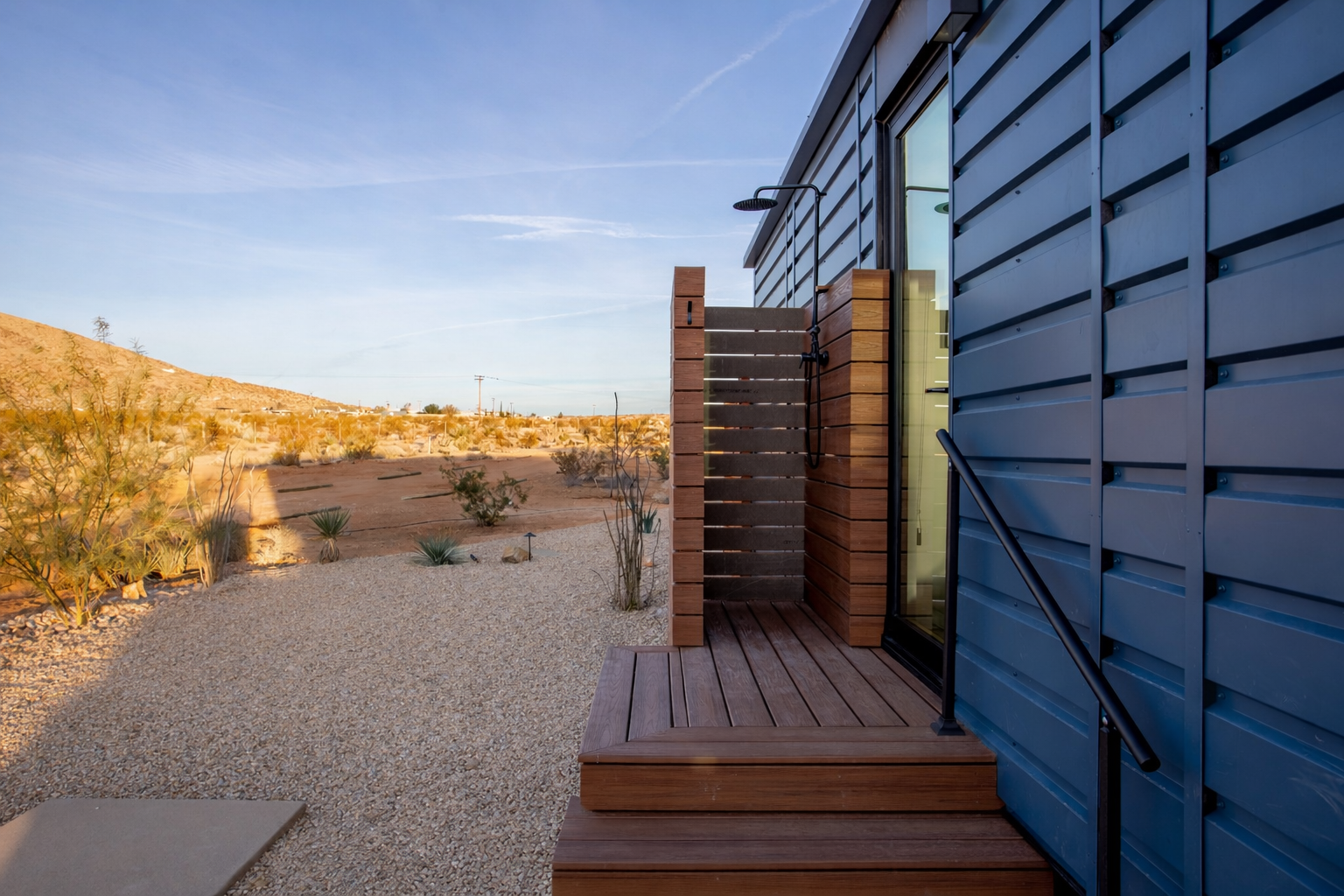 An outdoor shower with a wooden platform and blue metal siding, set in a desert landscape with sparse desert plants and a distant hill under a clear sky.