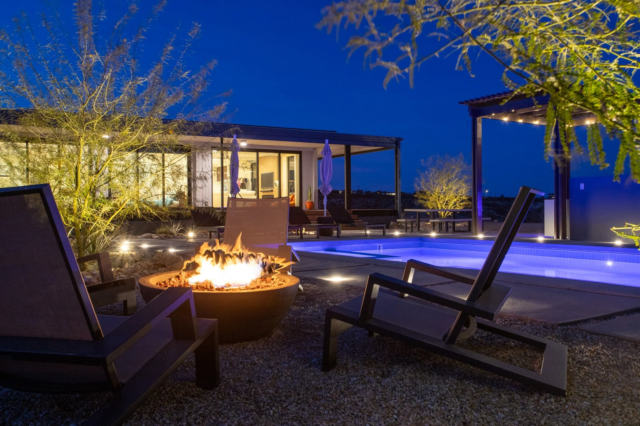 Night view of a modern backyard with a swimming pool illuminated in blue, surrounded by lounge chairs, a lit fire pit, and outdoor lighting, with a house visible in the background.