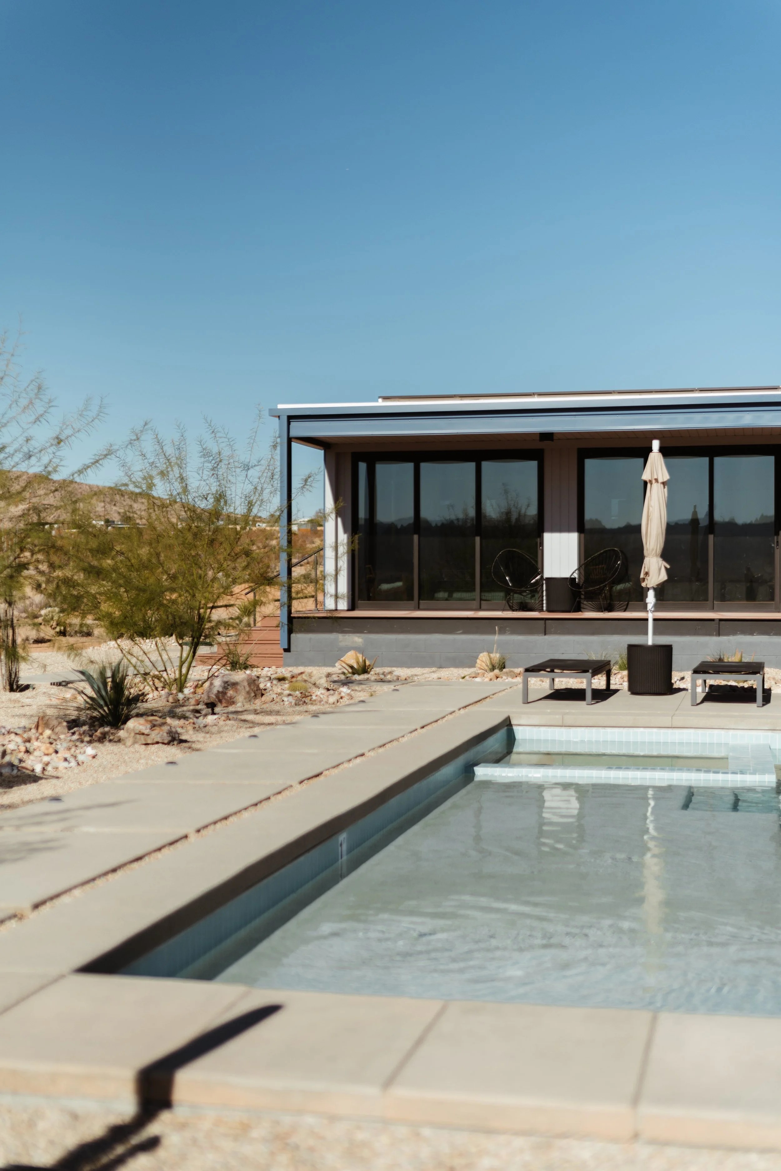 A modern house with large glass sliding doors, a swimming pool, and desert landscaping under a clear blue sky.