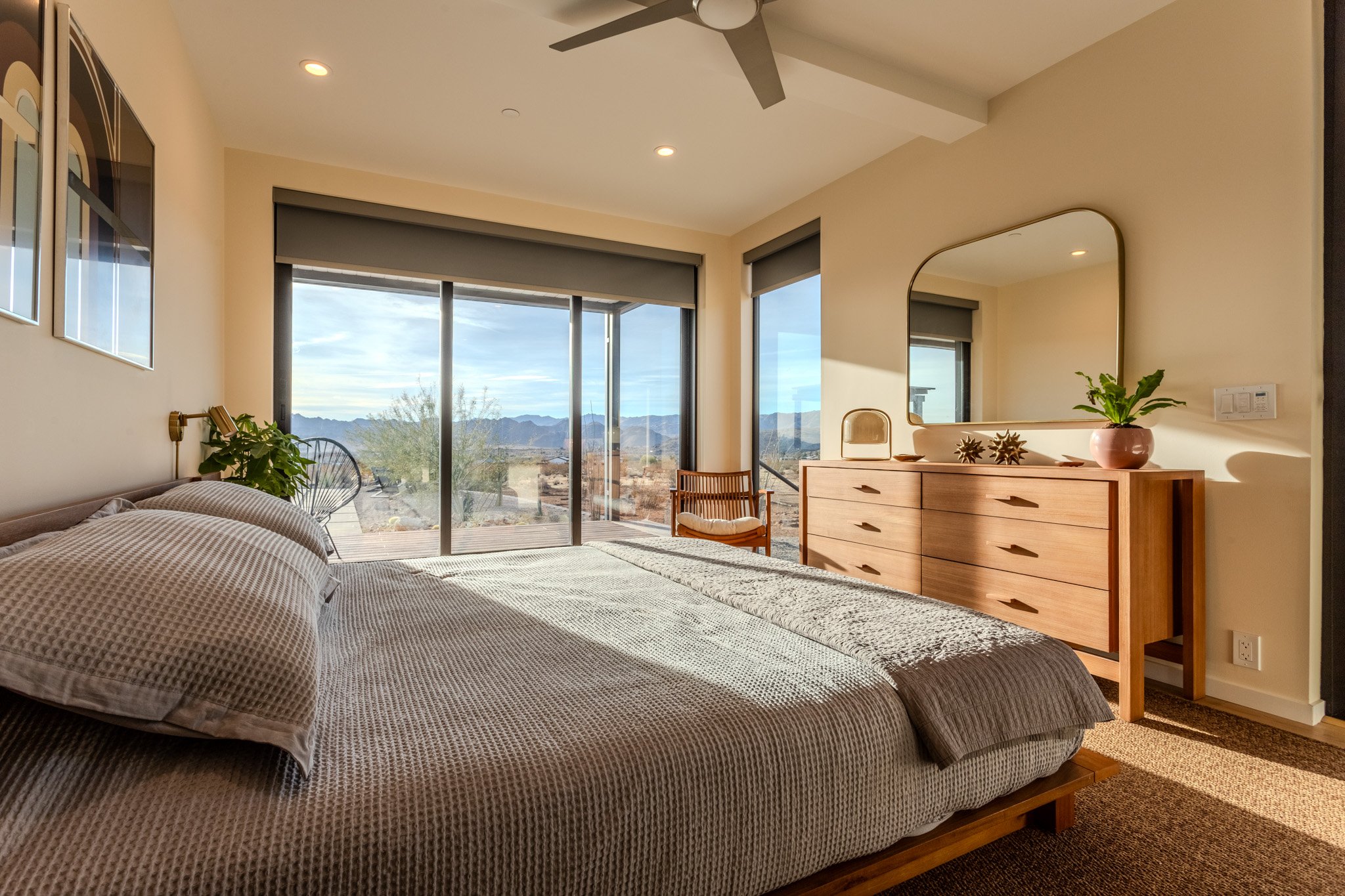 A bedroom with a large bed, a wooden dresser with a mirror, a chair, two potted plants, and a view through sliding glass doors of desert mountains and sky.