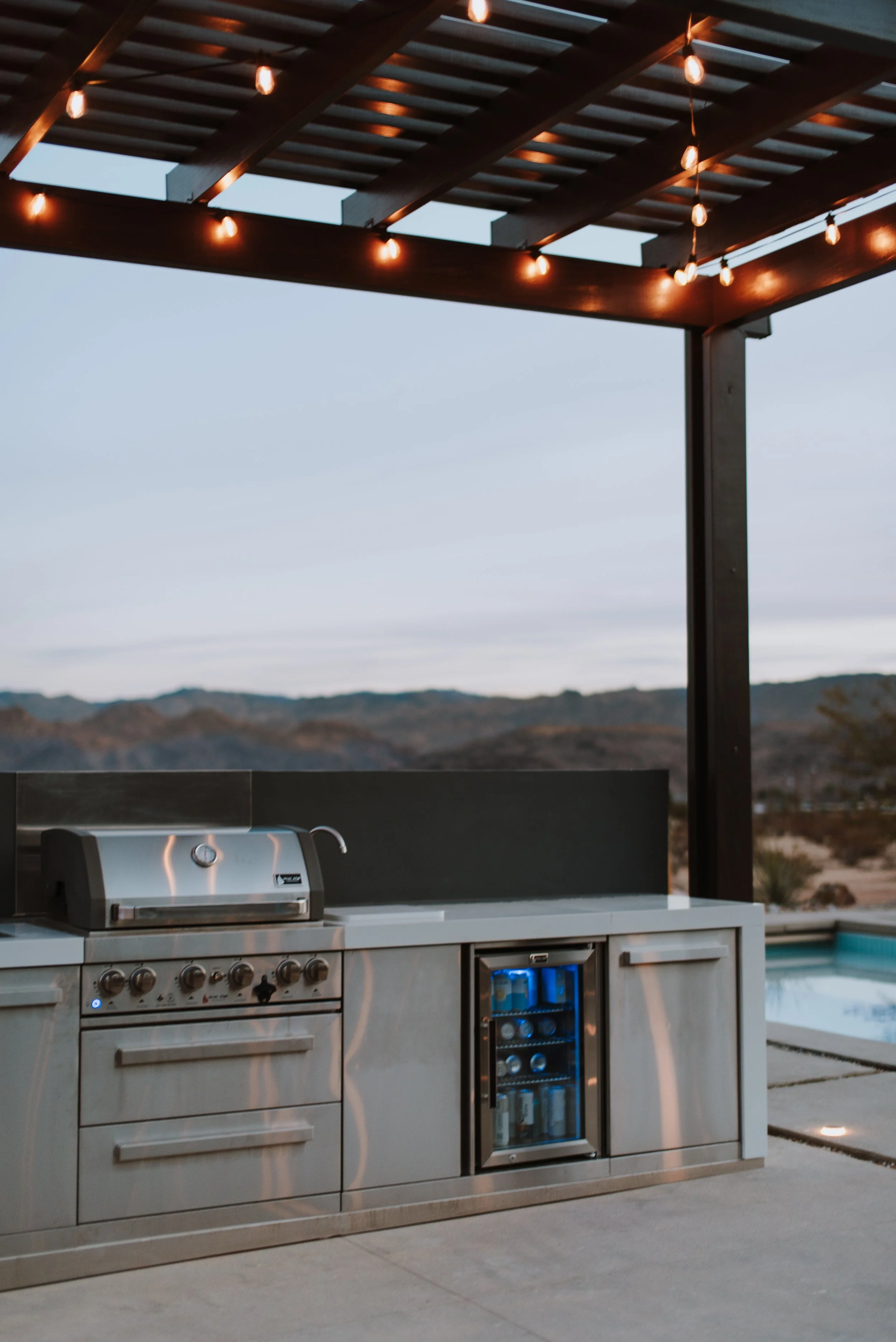 Outdoor kitchen with a stainless steel grill and mini fridge under a wooden pergola with string lights, overlooking a desert landscape.
