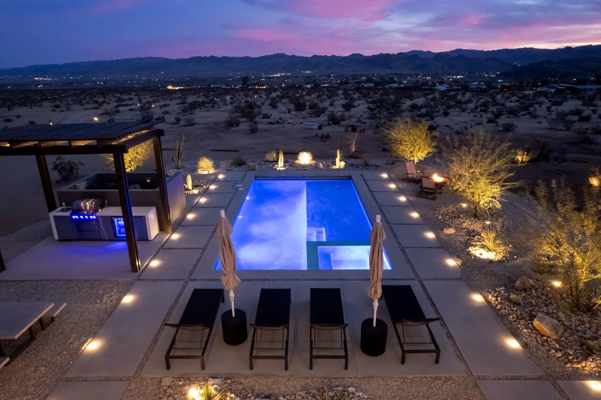 Night view of a luxurious backyard pool with surrounding lounge chairs, umbrellas, and outdoor seating, overlooking a desert landscape with mountains in the distance and a colorful sunset sky.