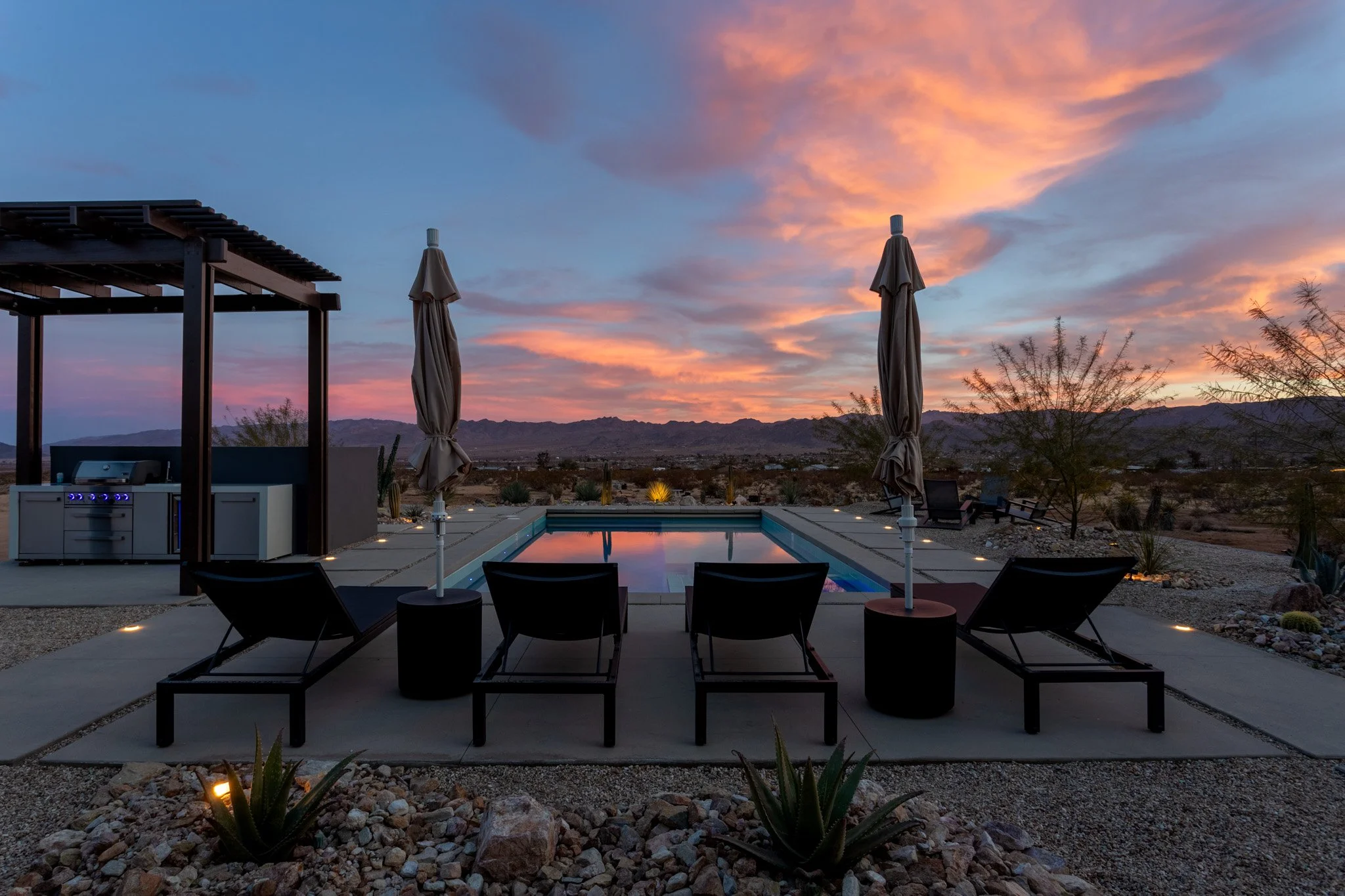 Outdoor swimming pool with lounge chairs, umbrellas, and a barbecue area at sunset in a desert landscape.