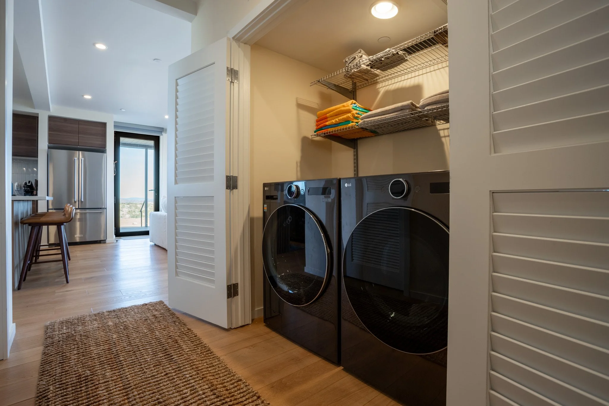 A laundry room with a black front-load washer and dryer, open shelving with folded towels, and louvered closet doors, with a view of a modern kitchen and balcony in the background.