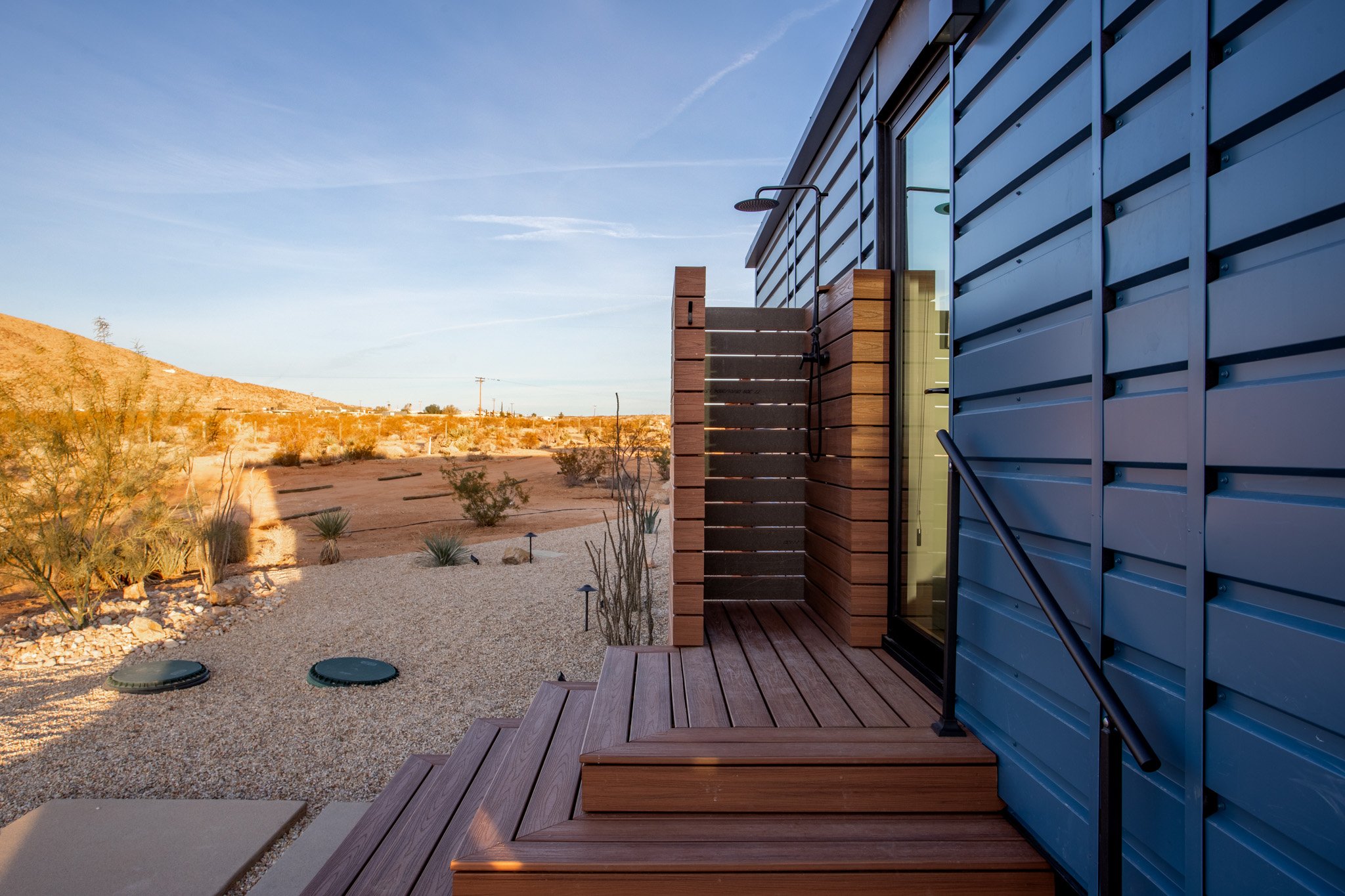 An outdoor shower with a wooden platform and blue metal siding, set in a desert landscape with sparse desert plants and a distant hill under a clear sky.