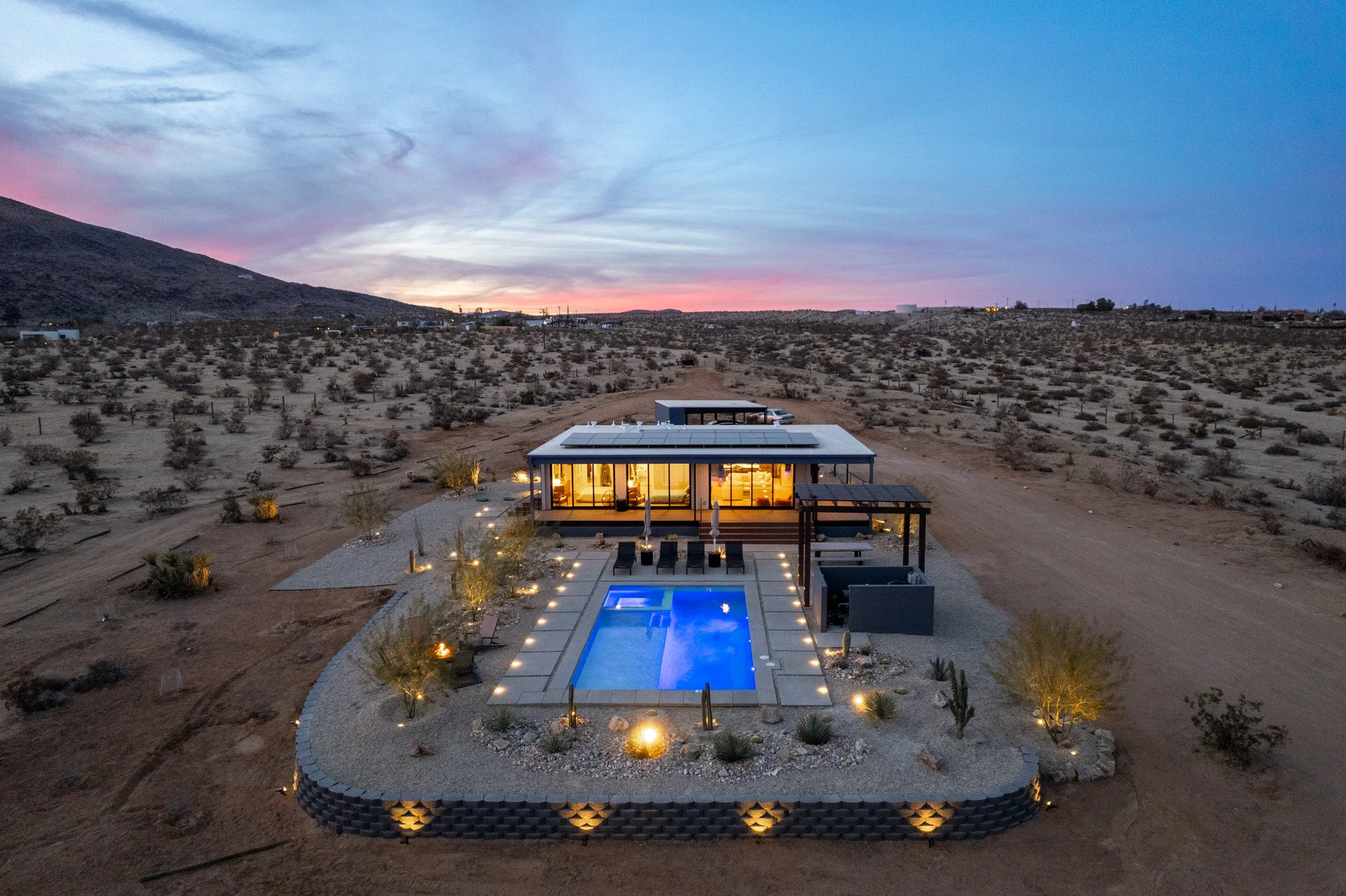 A modern house with large glass windows, illuminated from inside, surrounded by desert landscape with sparse shrubs and a mountain, at sunset. The house features an outdoor pool lit with blue lights, with poolside chairs and a small shaded lounging a