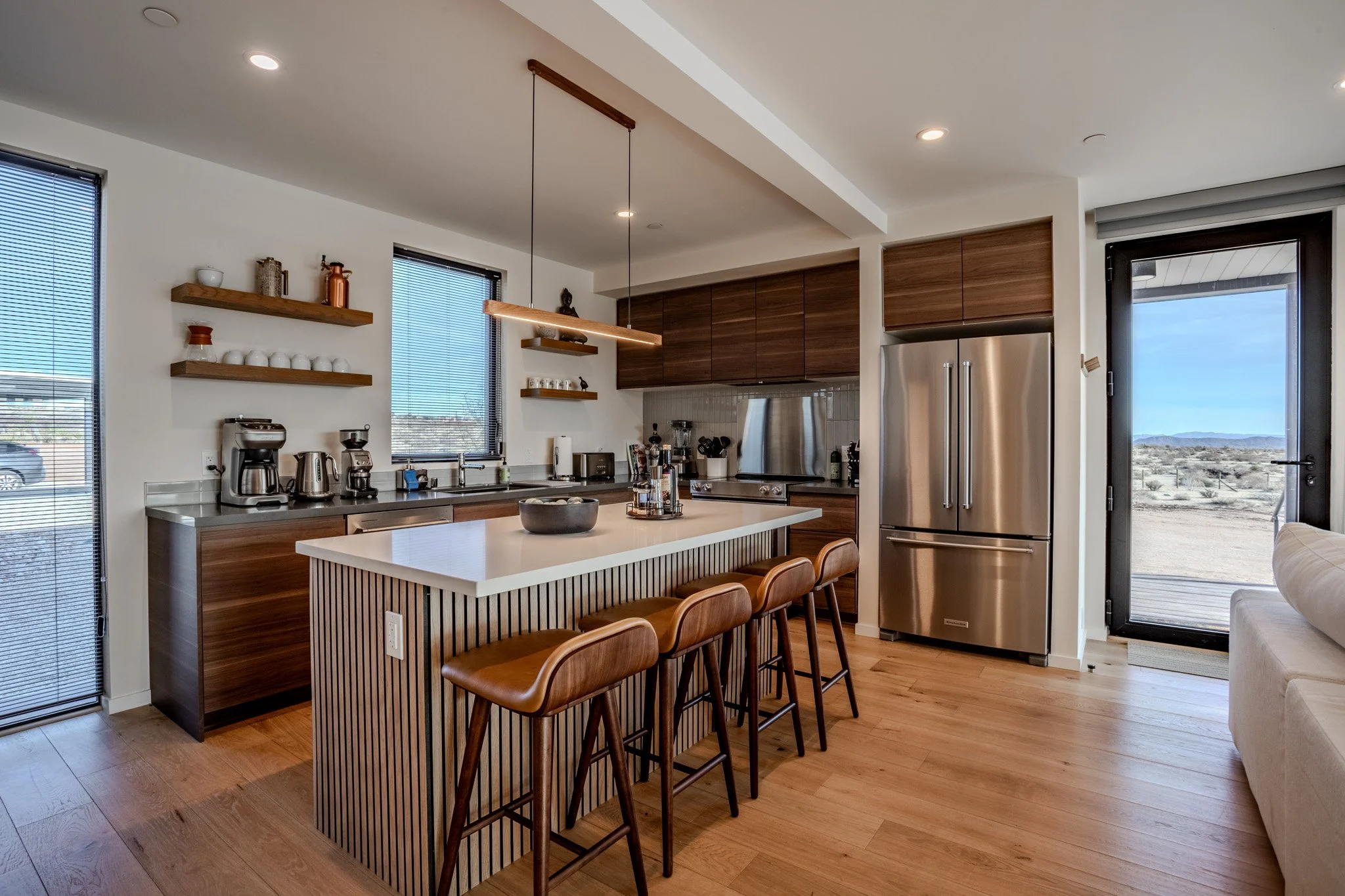 Modern kitchen with wooden cabinets, stainless steel appliances, a central island with four brown leather chairs, open shelves with dishes and decor, and a view of a desert landscape through a glass door.