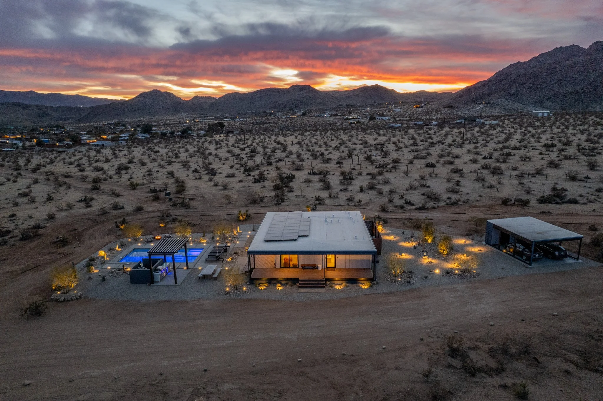 A modern house with a flat roof, illuminated at dusk in a desert landscape. The house features solar panels on the roof, a porch, and large windows. Surrounding the house are small trees with outdoor lighting, a swimming pool with a shaded seating ar
