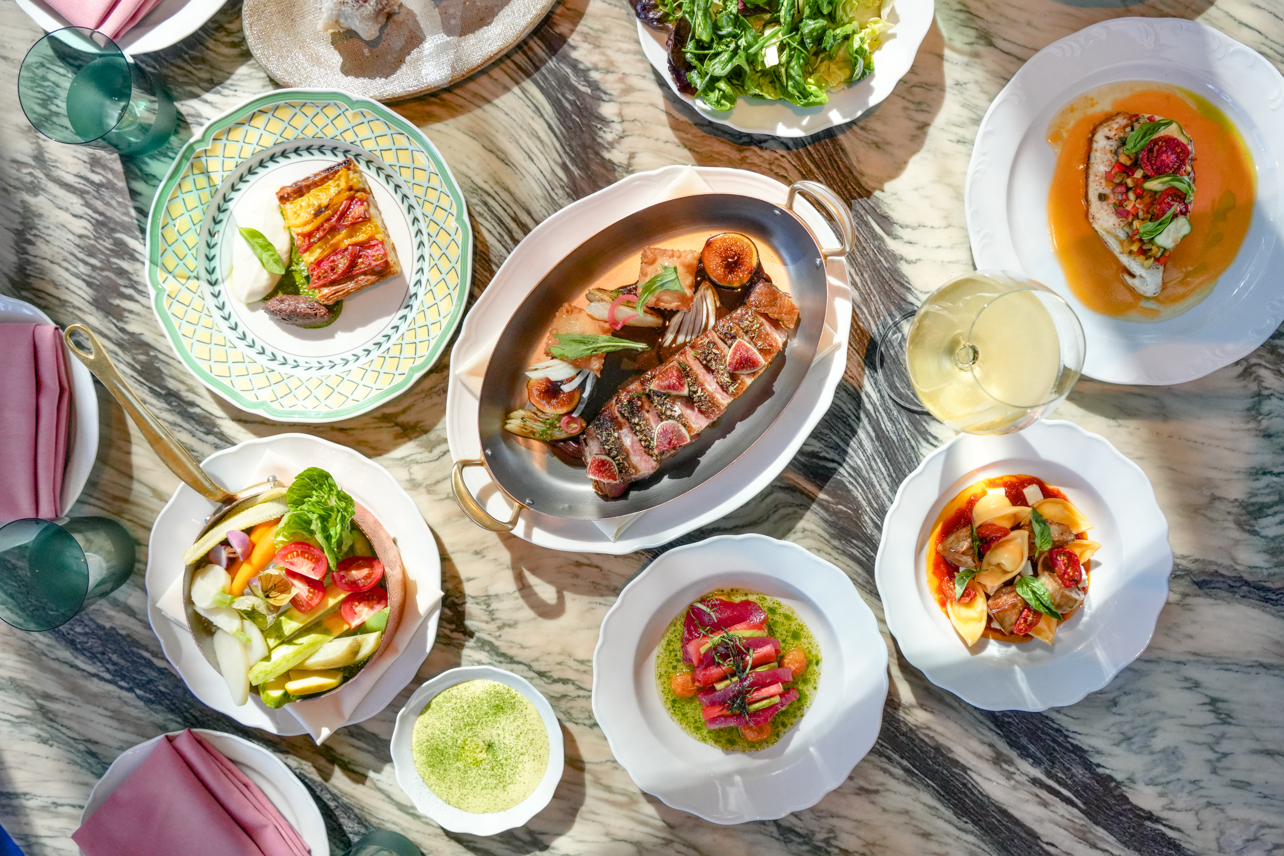 A variety of colorful dishes including salads, grilled meats, and sauces are displayed on a marble table, with glasses of wine and water.