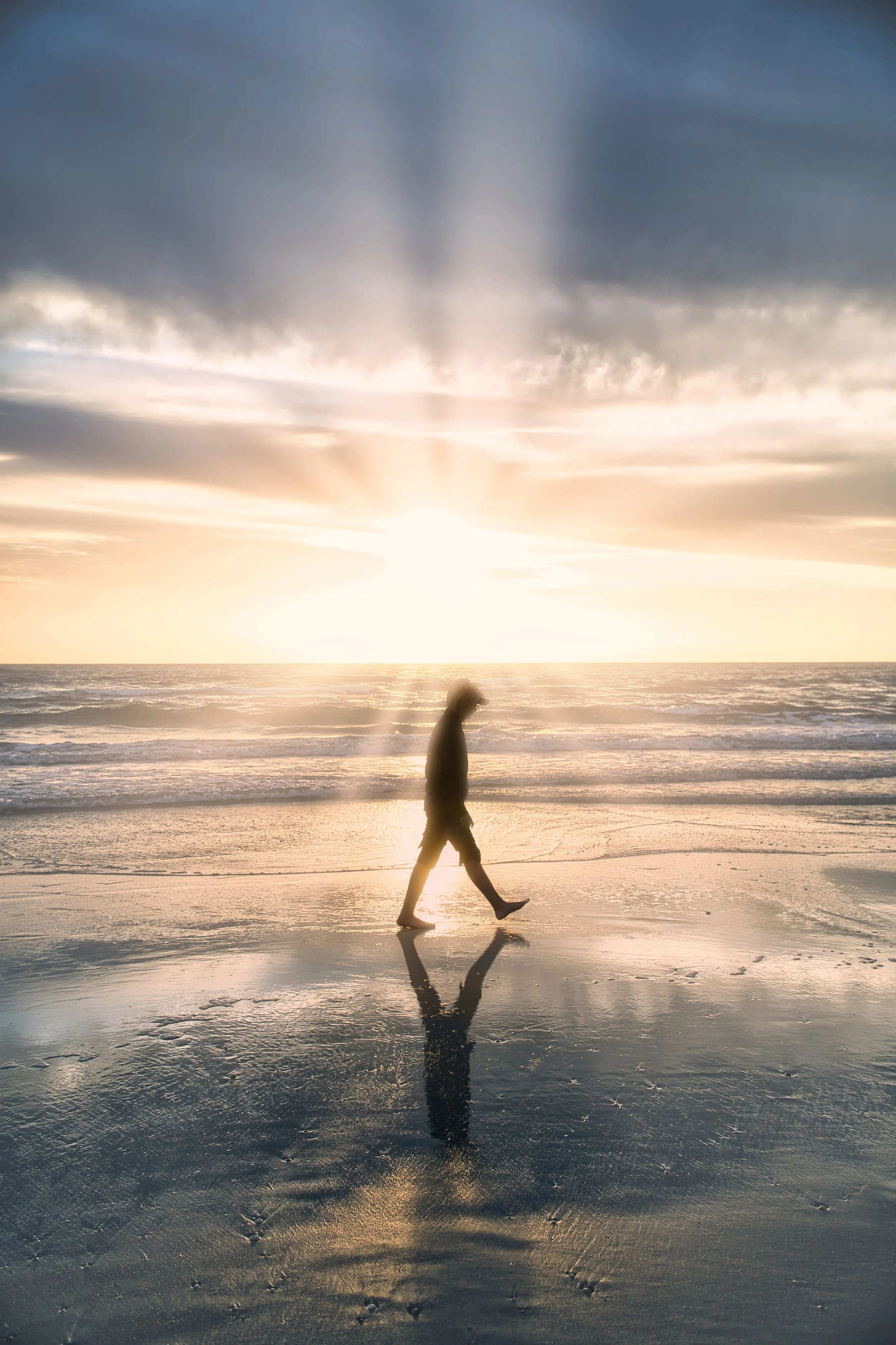 Eine Person läuft am Strand während des Sonnenuntergangs, mit reflektierendem Wasser auf dem Boden, Himmel mit Wolken und Sonnenstrahlen im Hintergrund.