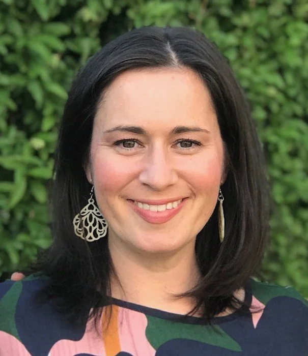 A woman with shoulder-length black hair, light skin, and blue eyes smiling outdoors in front of green foliage, wearing large earrings and a colorful top.