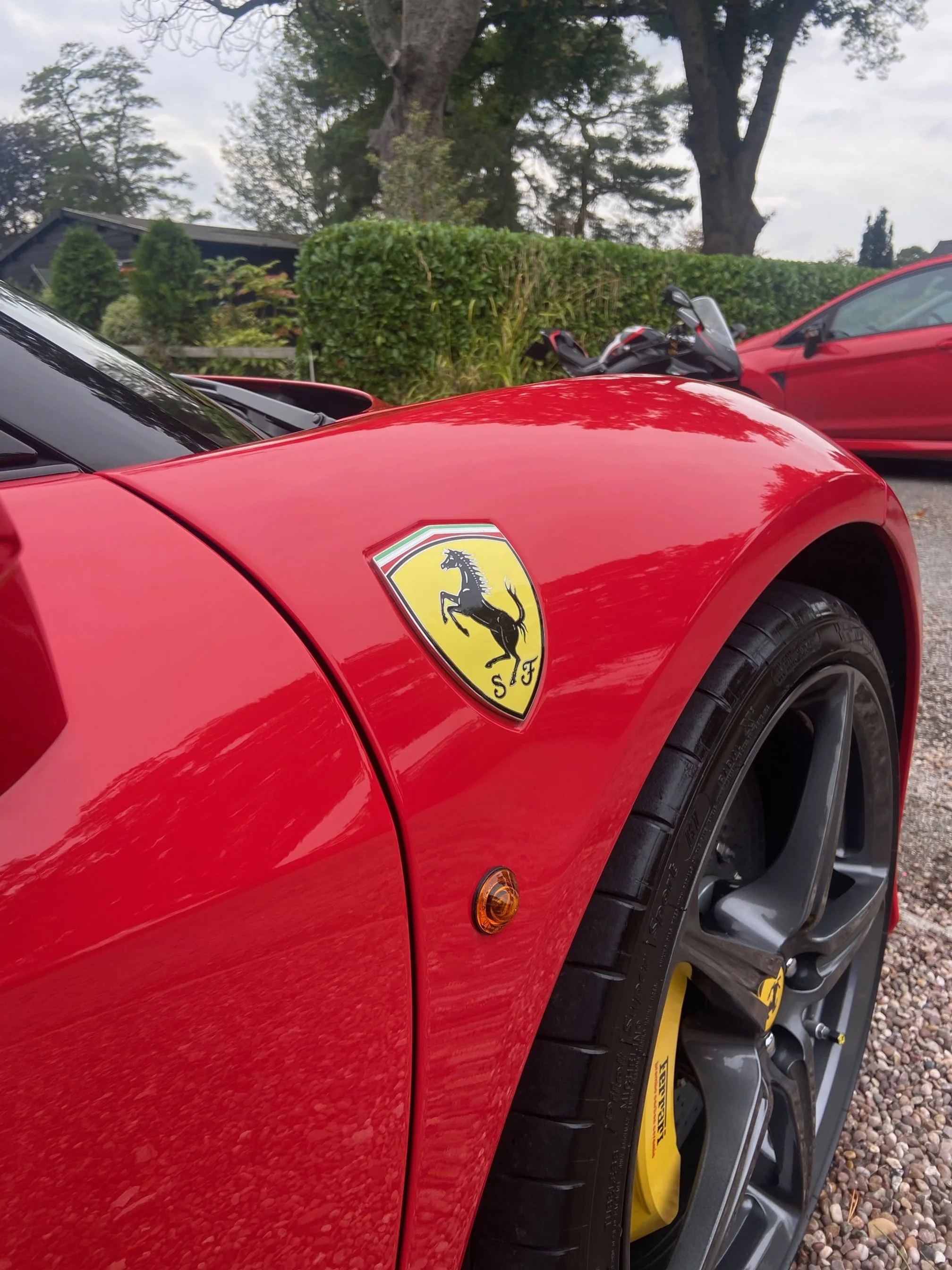 Close-up of a red Ferrari sports car with the Ferrari emblem on the front fender, showing part of the wheel and tire against a background of trees, bushes, and other vehicles.