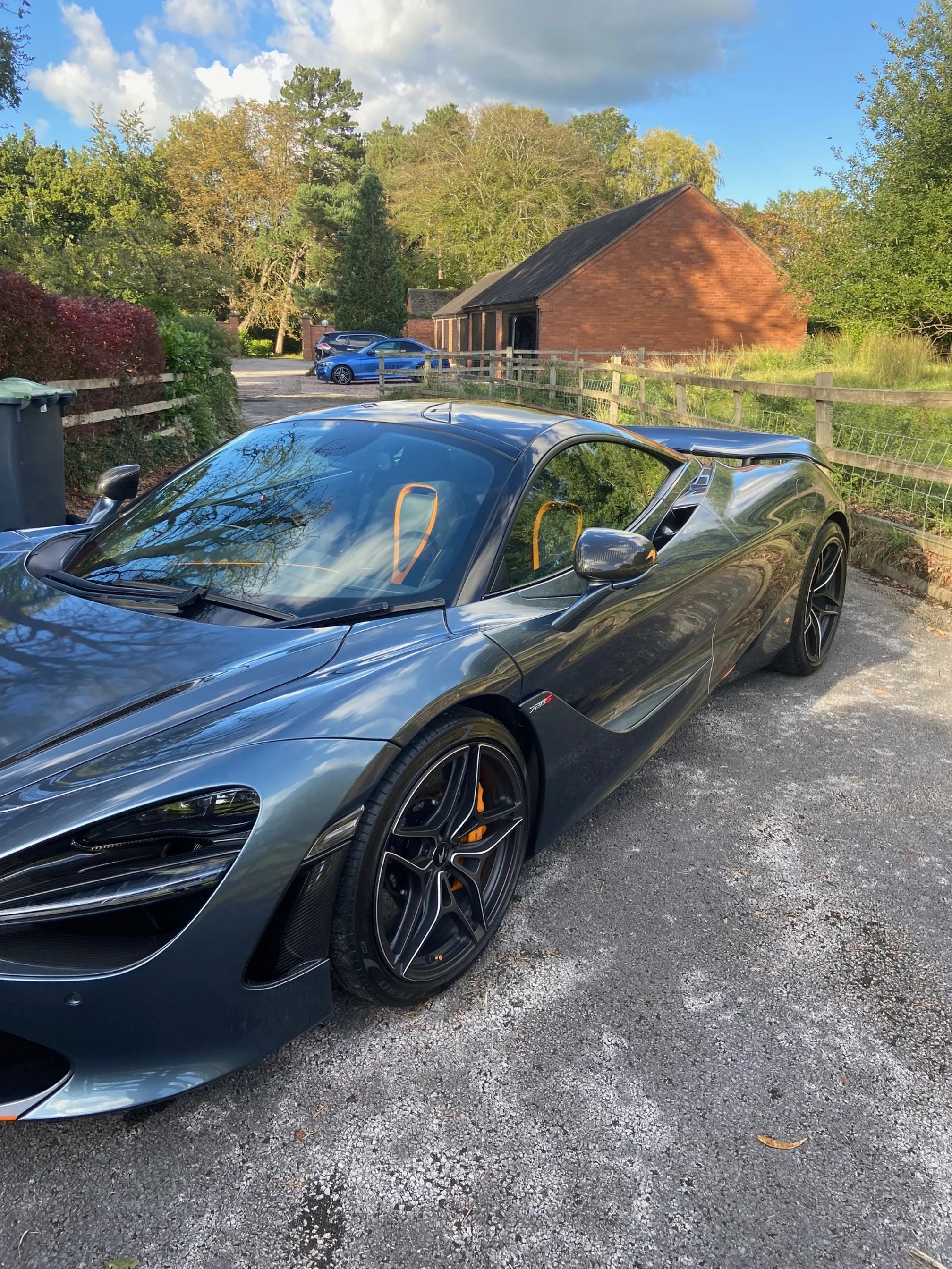 A sleek, dark gray sports car parked on a gravel driveway with a scenic background of trees, a wooden fence, a small brick building, and a blue sky with some clouds.