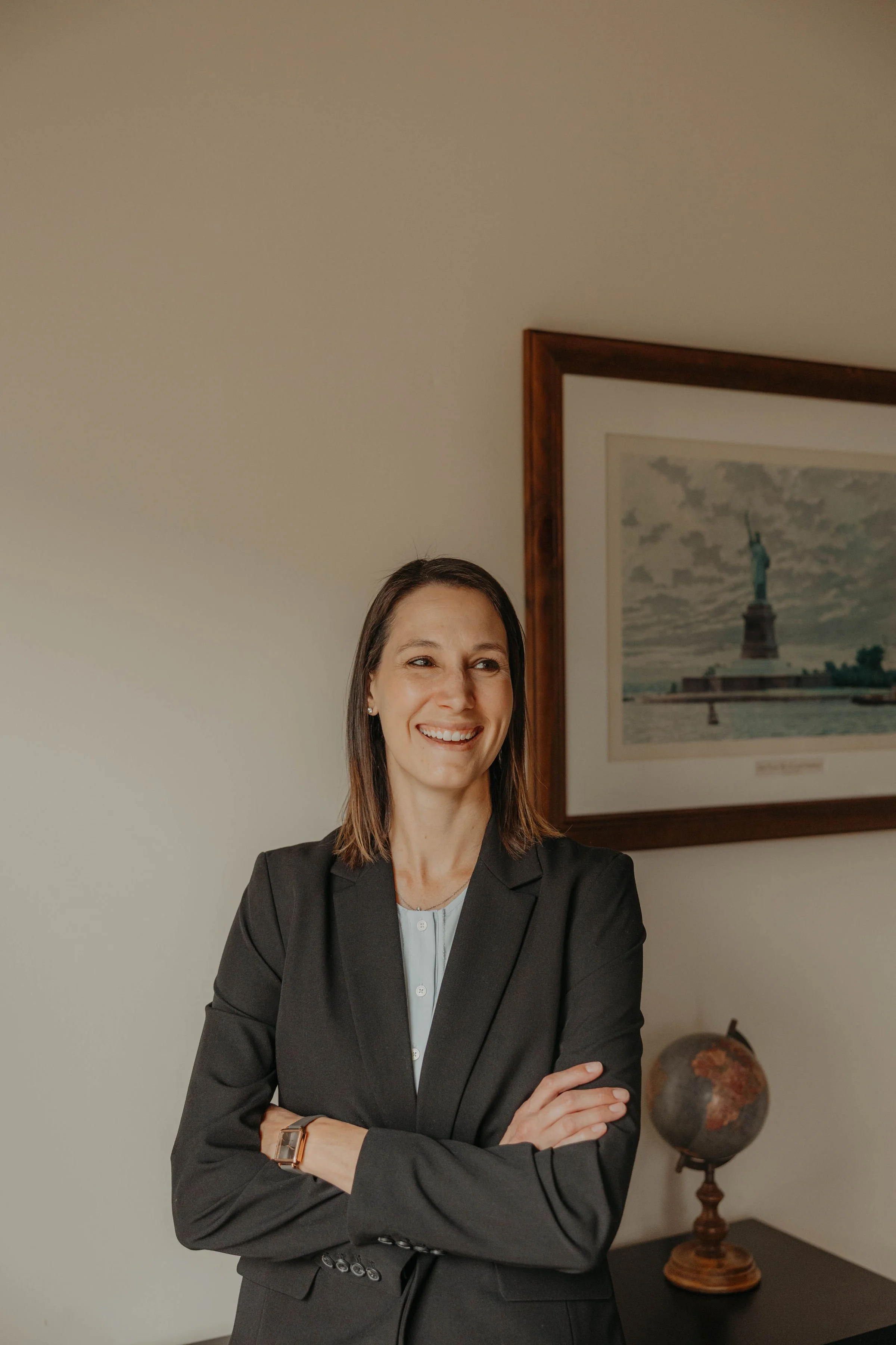 A smiling woman in a black blazer standing with arms crossed in an office or study, with a framed picture of the Statue of Liberty on the wall and a globe on a table in the background.