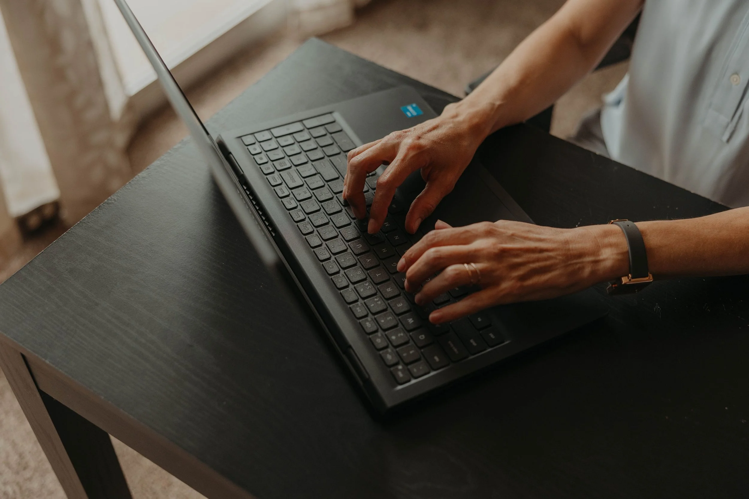 Person using a laptop on a dark wooden desk, typing on the keyboard with a watch and ring on their hands.