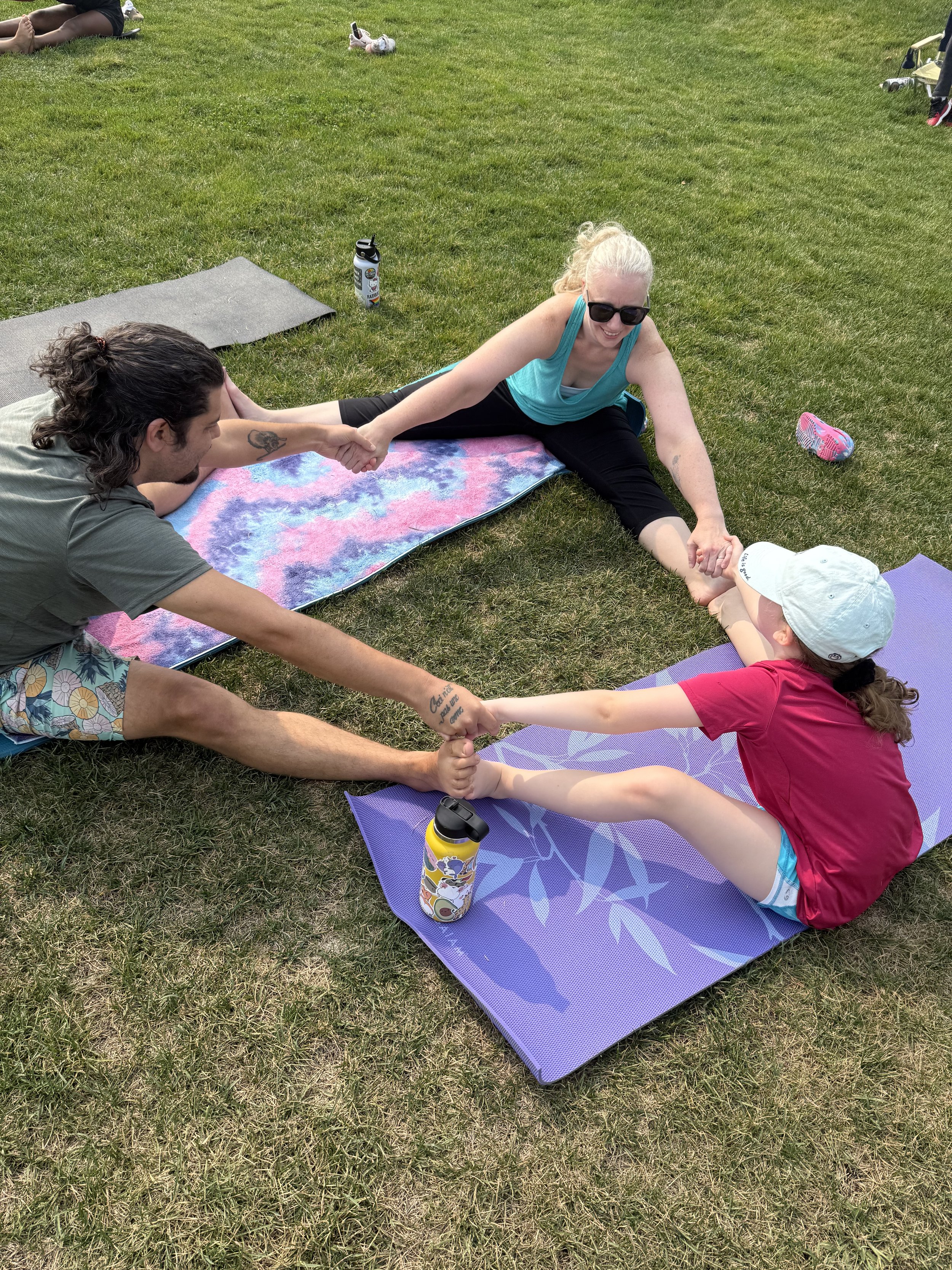 A woman and a girl are doing a stretching exercise on yoga mats outdoors on grass. The woman is wearing sunglasses and black pants, while the girl is in a red shirt and white hat. They are holding hands with another person who is out of frame. Water bottles are nearby, and there are more mats and people in the background.