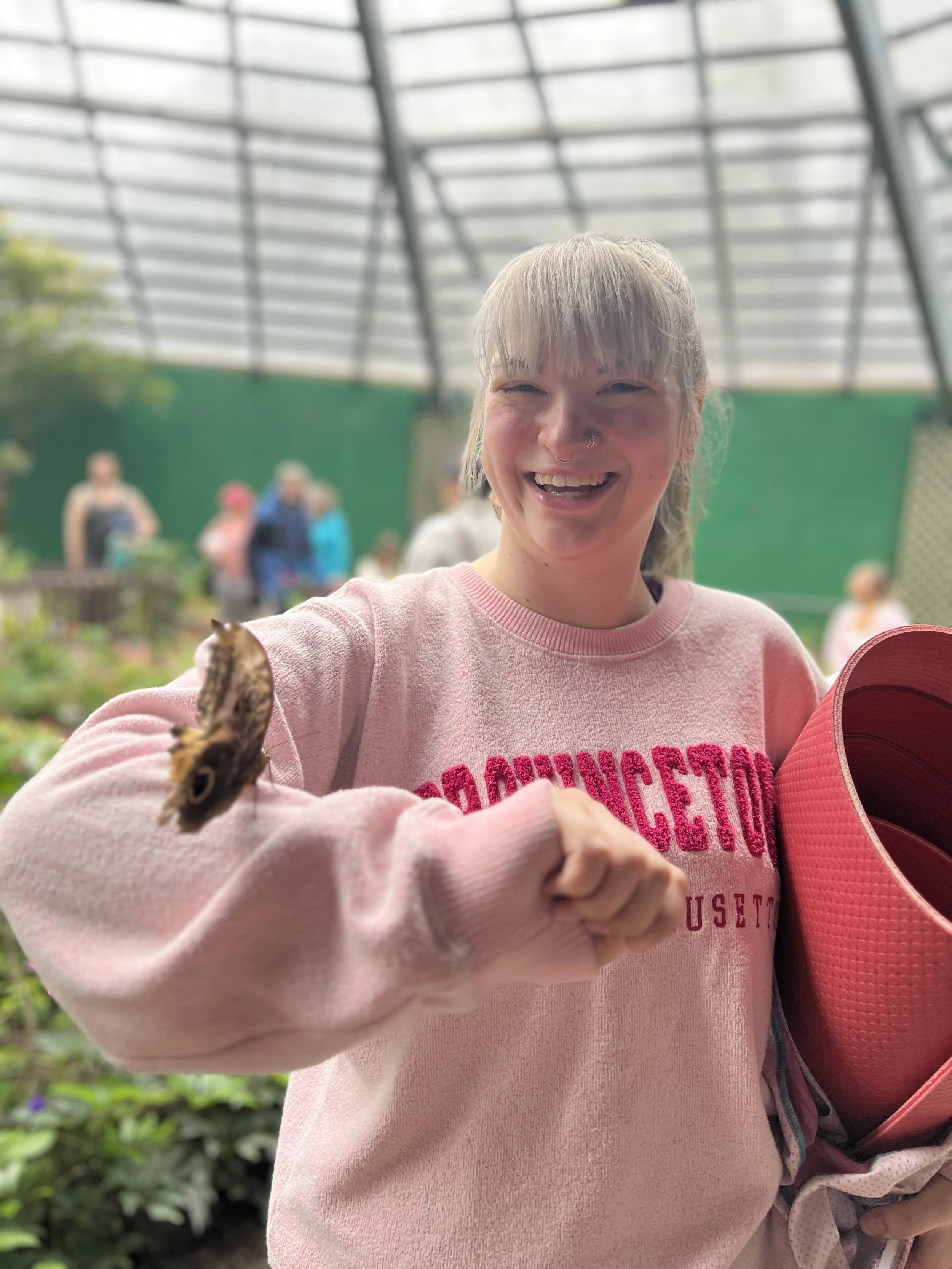 A smiling person with light colored hair and a nose piercing, wearing a pink sweatshirt, holding a colorful butterfly-shaped object, inside a greenhouse or similar structure, with blurred people and greenery in the background.