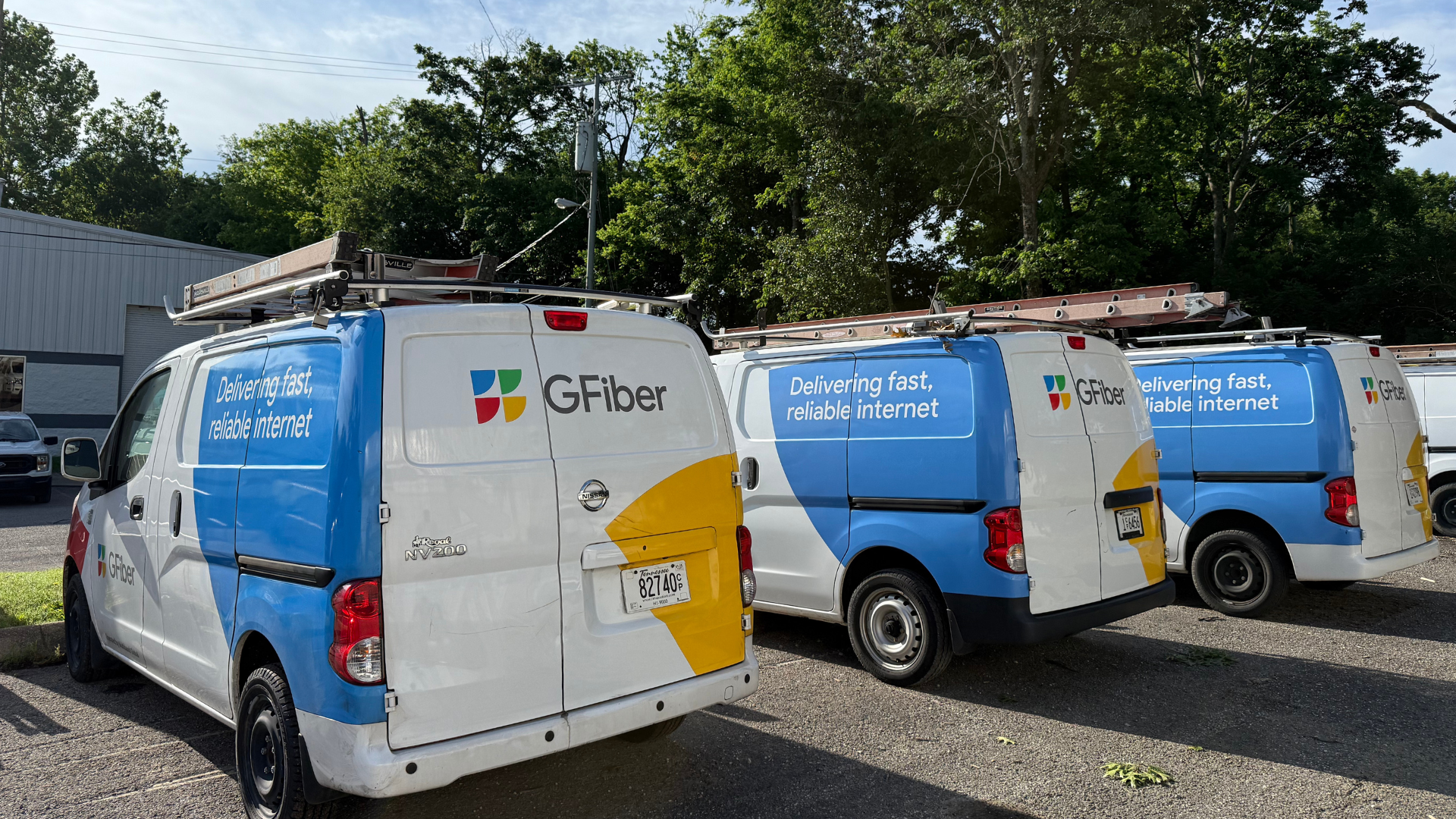Three GFiber delivery vans parked outdoors near a building with trees in the background; the vans are white and blue with the GFiber logo and slogan 'Delivering fast, reliable internet'.
