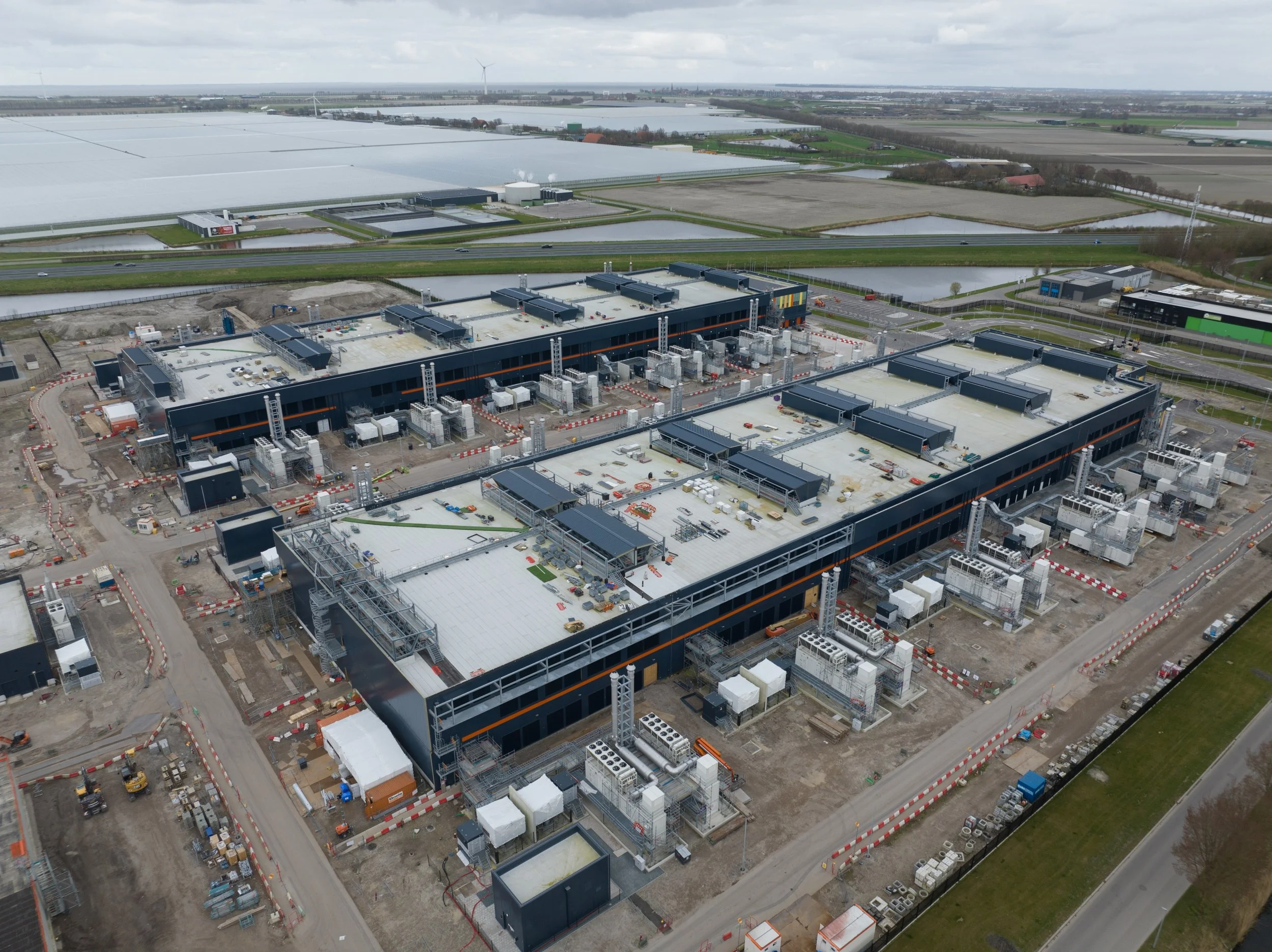 Aerial view of a large construction site with two modern, rectangular buildings in progress, surrounded by construction materials, machinery, and roads, with agricultural fields and water bodies in the background.