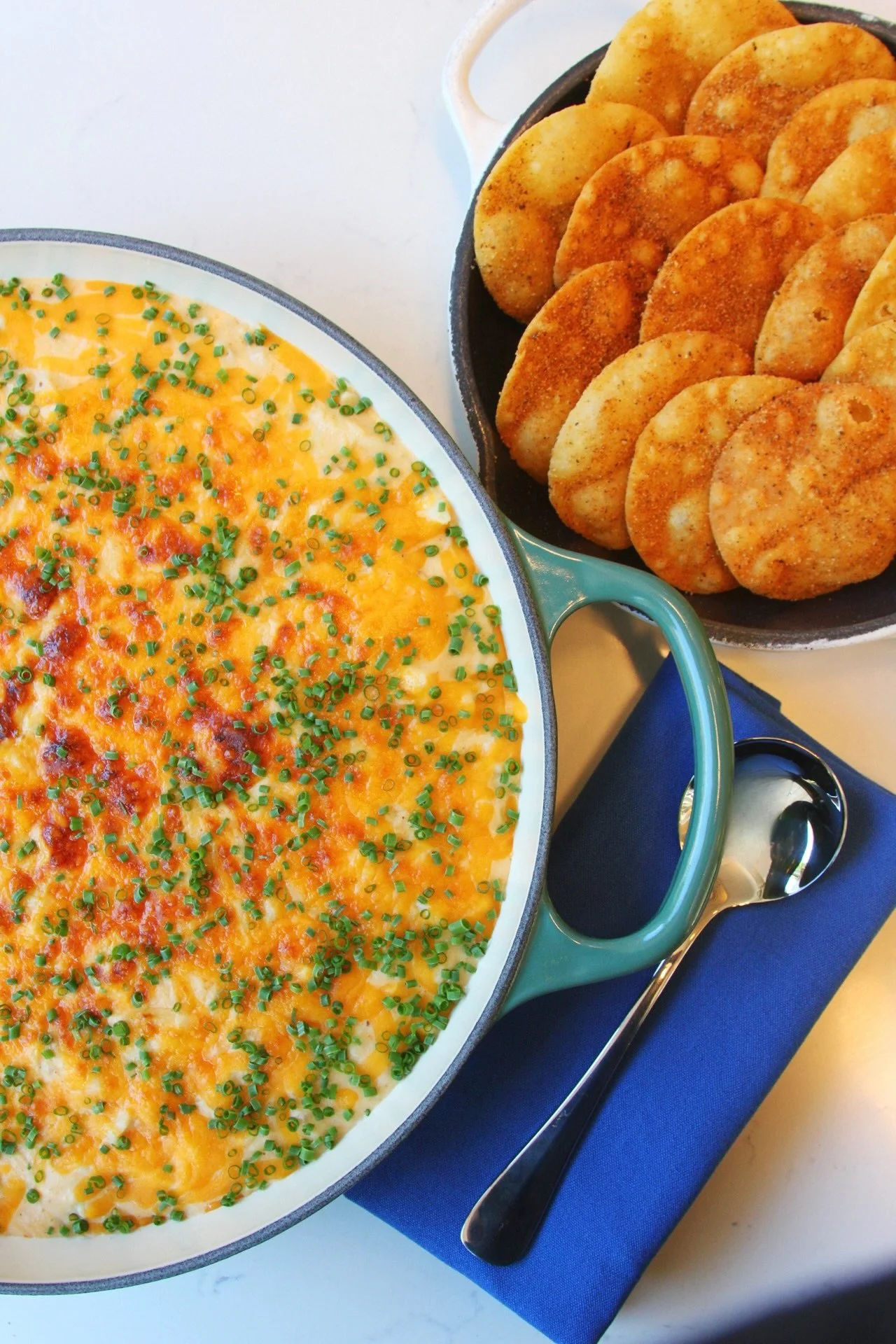 Baked dish topped with melted cheese and chopped green onions next to a bowl of fried potato pancakes with a spoon on a blue napkin.