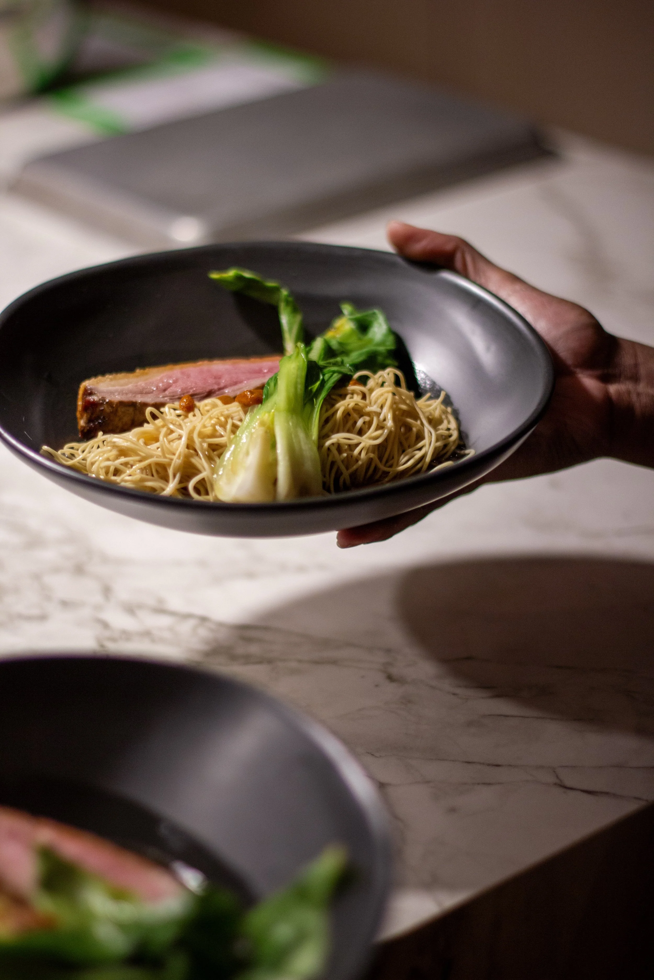 A black bowl of ramen with noodles, a green leafy vegetable, and a slice of pink beef, held by a person's hand over a white marble surface.