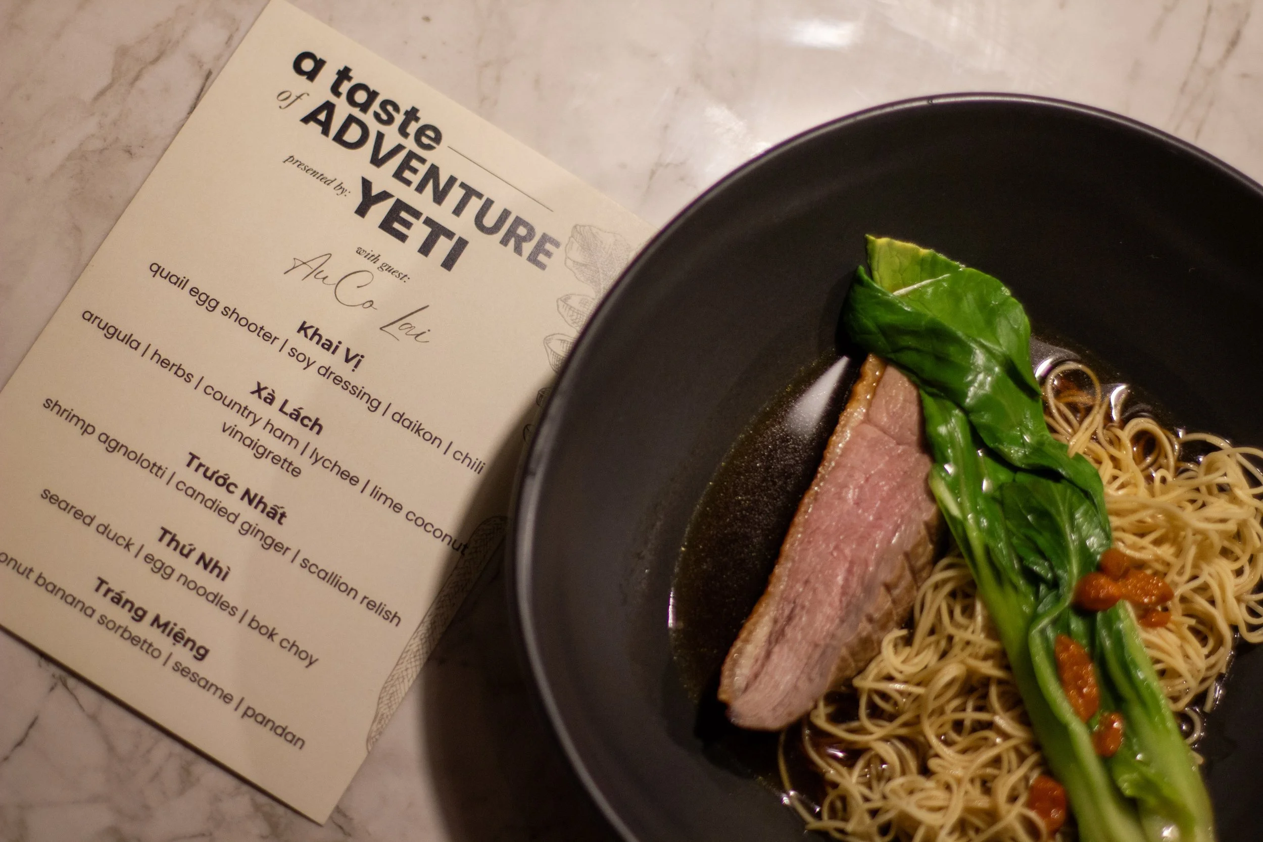 A black bowl containing noodles, a piece of cooked meat, fresh green bok choy, and small orange garnishes. Next to the bowl is a menu titled "a taste of ADVENTURE" presented by YETI with a guest chef, listed with Vietnamese dishes.