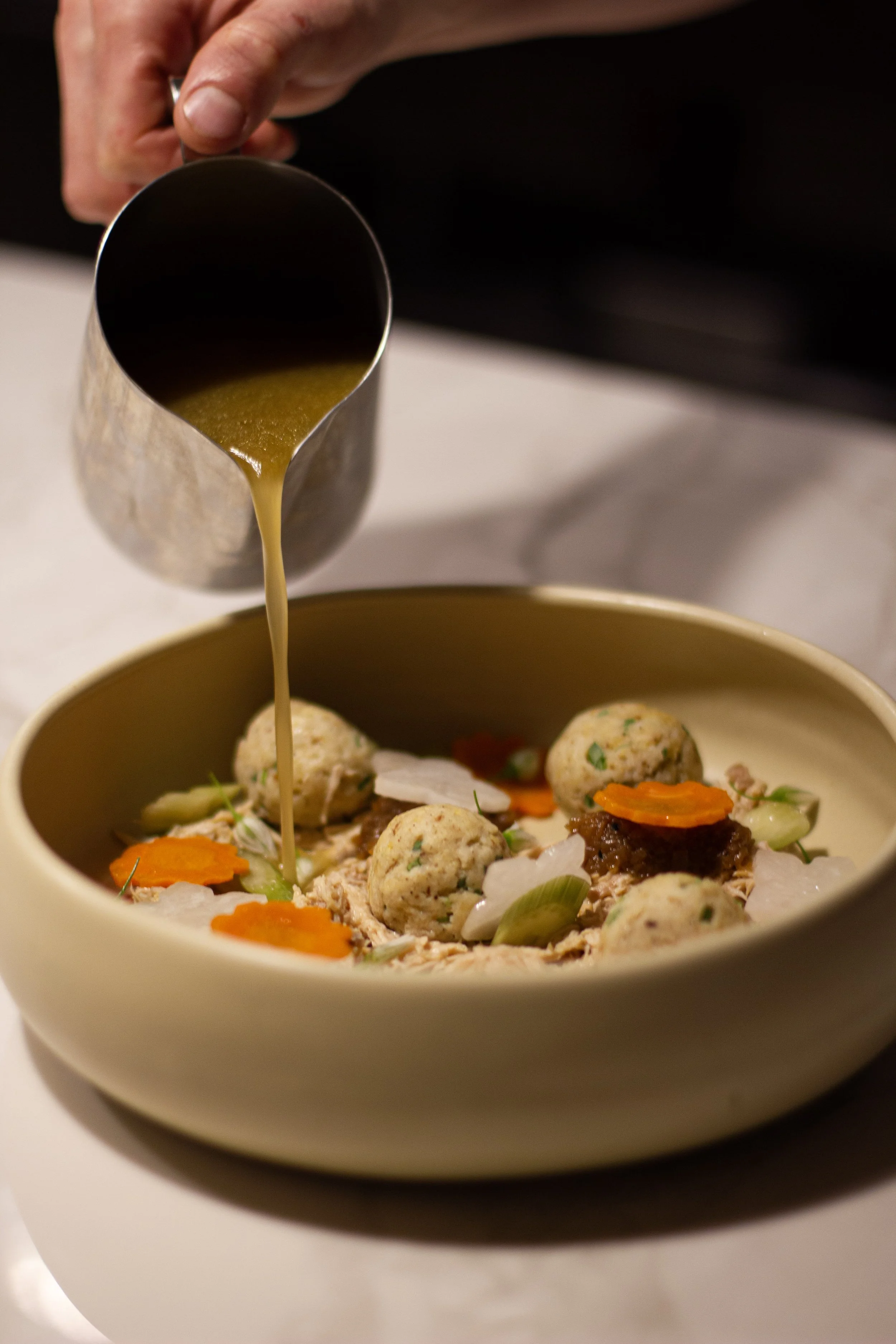 Chef pouring brown gravy or sauce over a dish with meatballs, sliced carrots, celery, and mashed potatoes in a beige bowl.